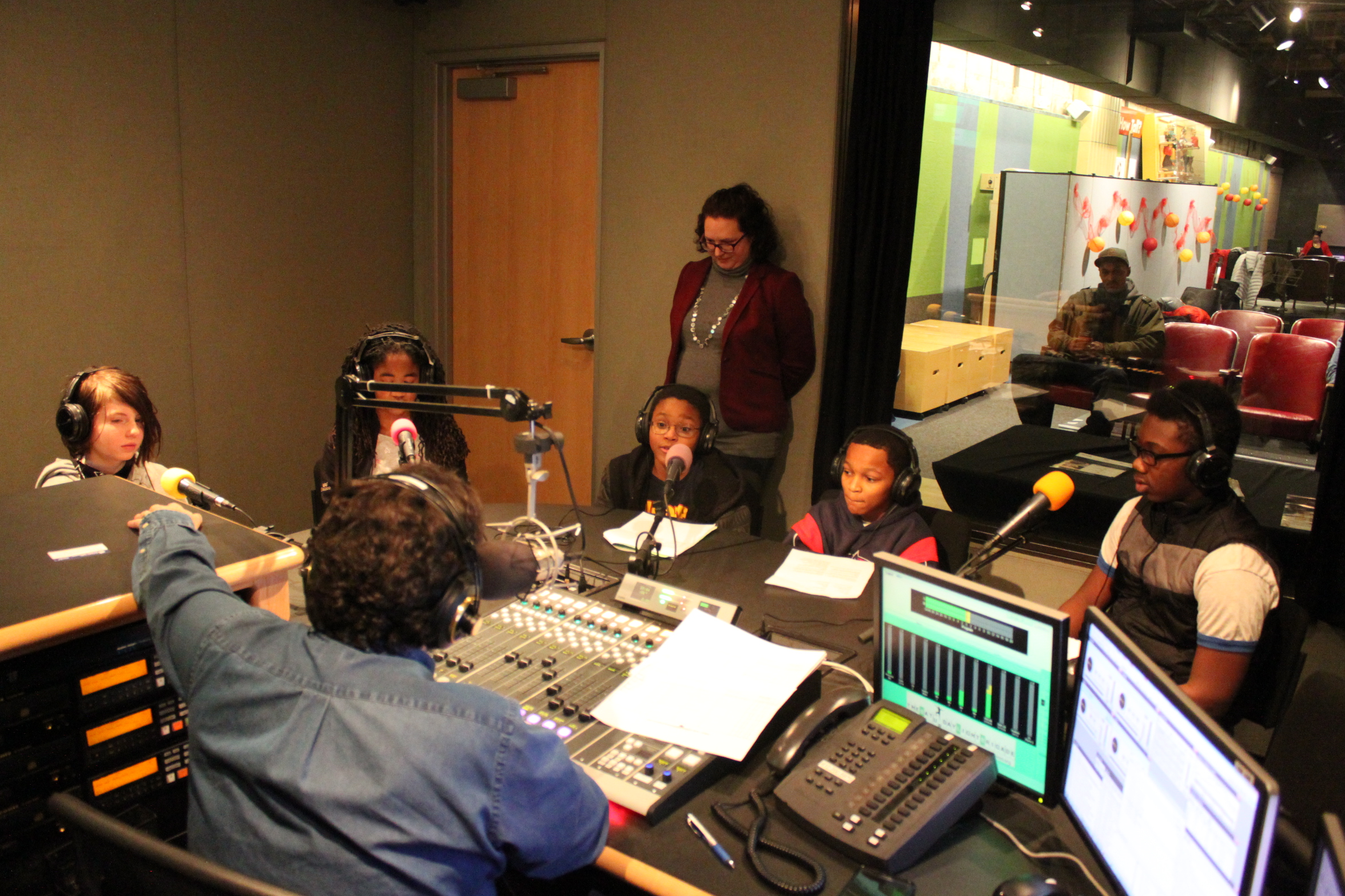 Five youth wearing headphones sitting behind microphones being interviewed by an adult wearing headphones sitting behind a microphone and a mixing board as an adult listens in the background in a radio studio