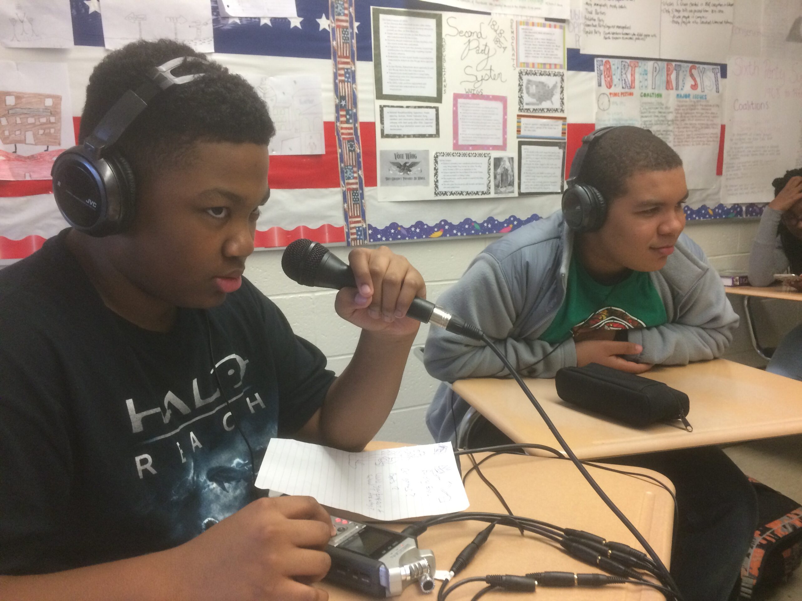 Two youth wearing headphones sitting in a classroom, one speaking into a microphone