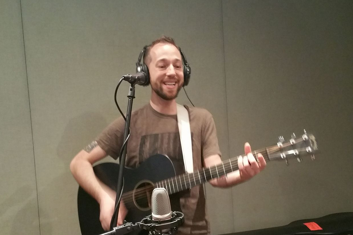 An adult wearing headphones playing acoustic guitar and singing in a radio studio