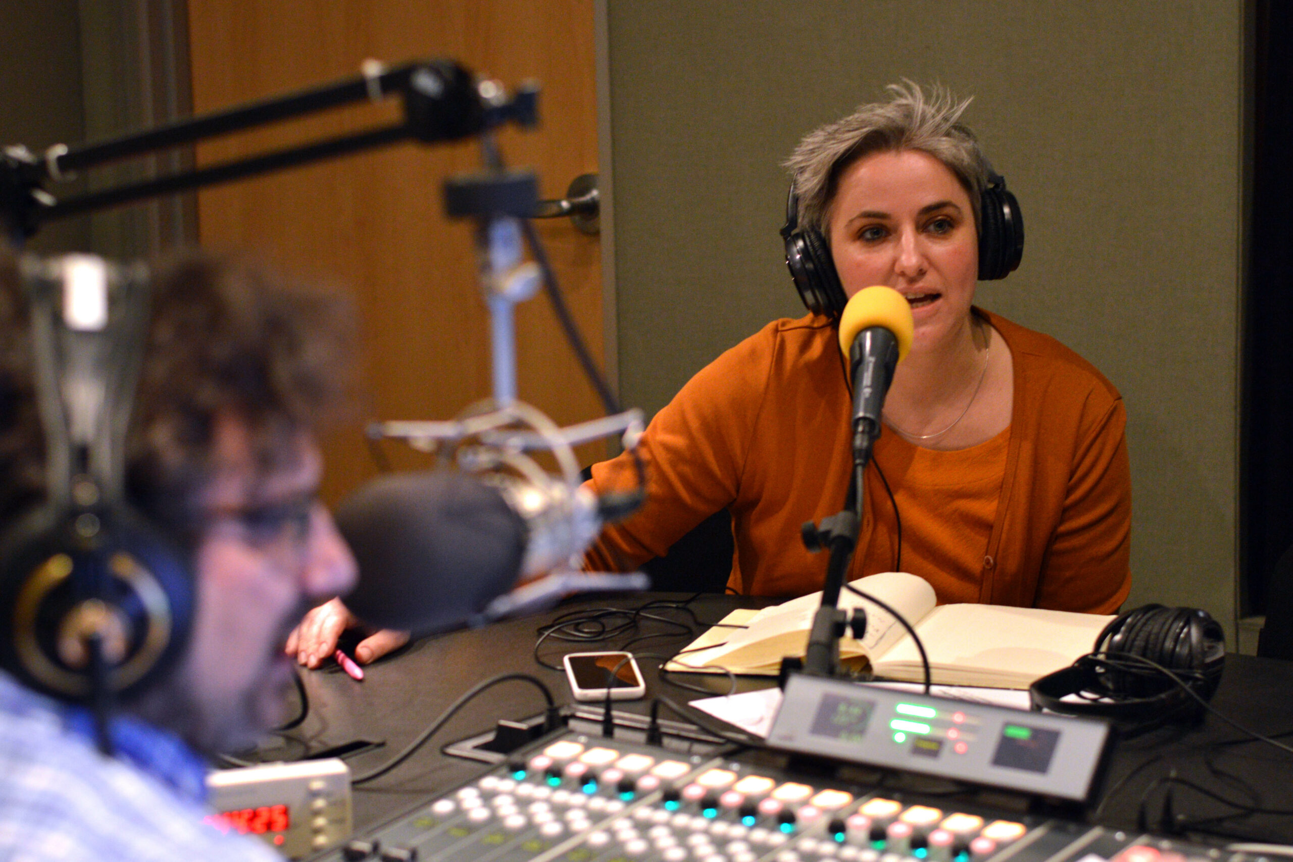 An adult wearing headphones speaking into a microphone being interviewed by an adult wearing headphones sitting behind a microphone and a mixing board in a radio studio