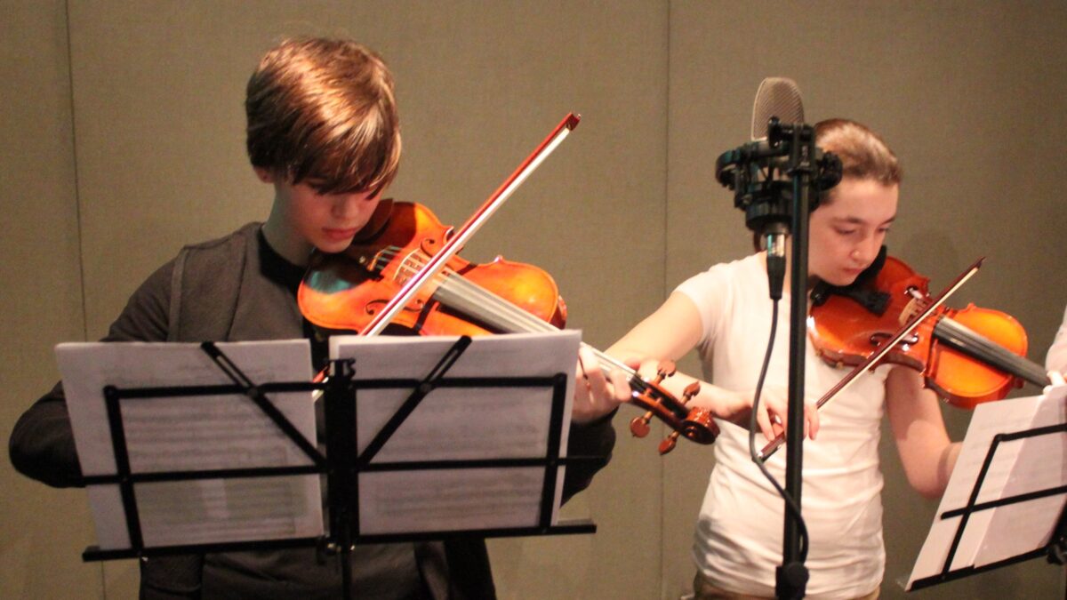 Two youth playing violins looking at sheet music on music stands in a radio studio