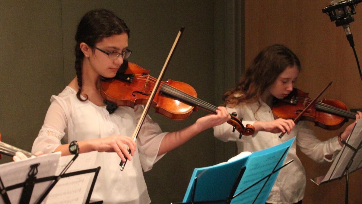 Two youth playing violins looking at sheet music on music stands in a radio studio