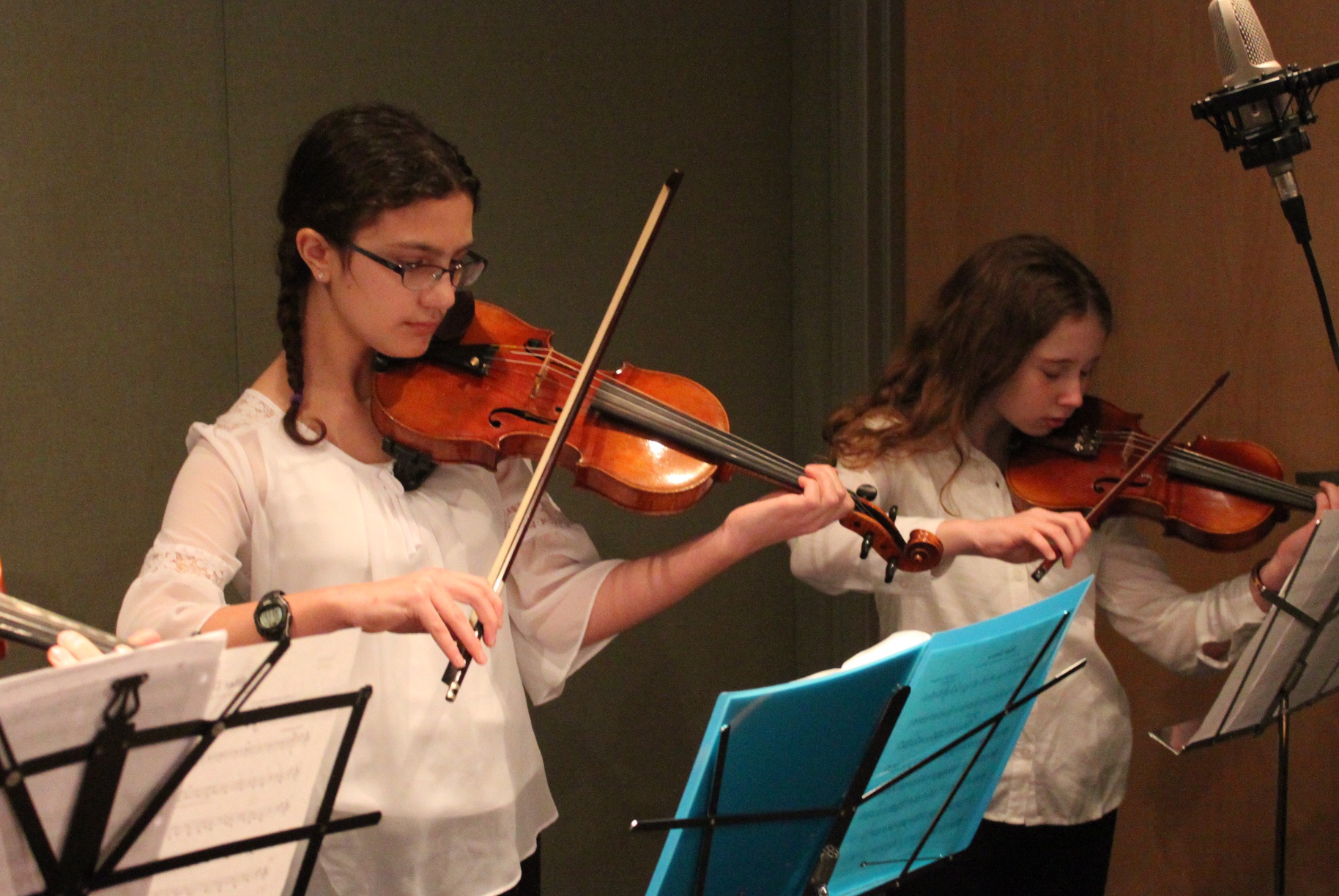 Two youth playing violins looking at sheet music on music stands in a radio studio