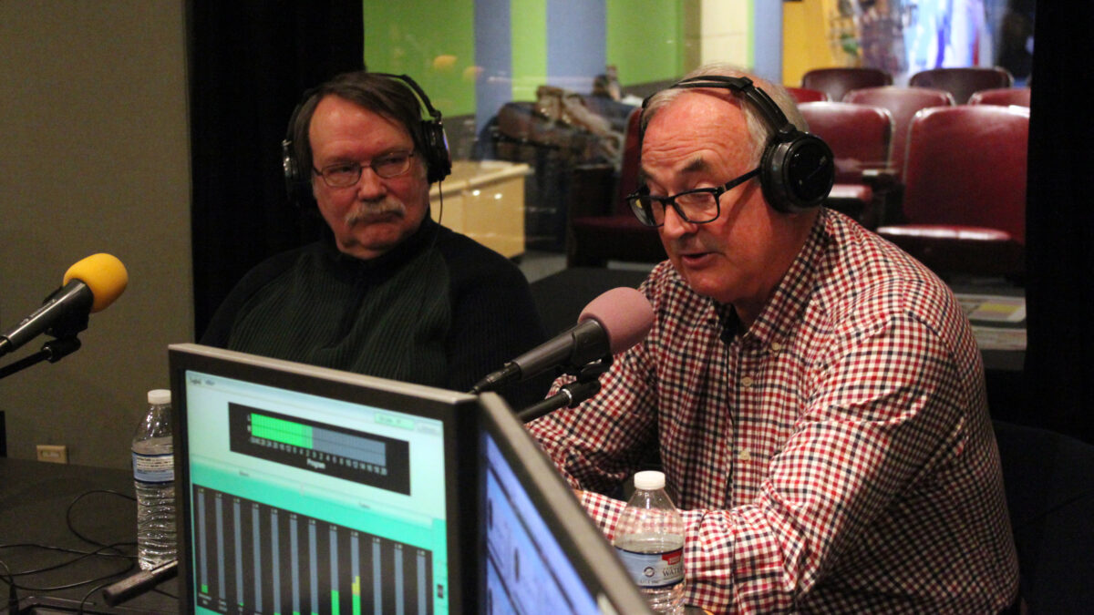 Two adults wearing headphones speaking into microphones in a radio studio.