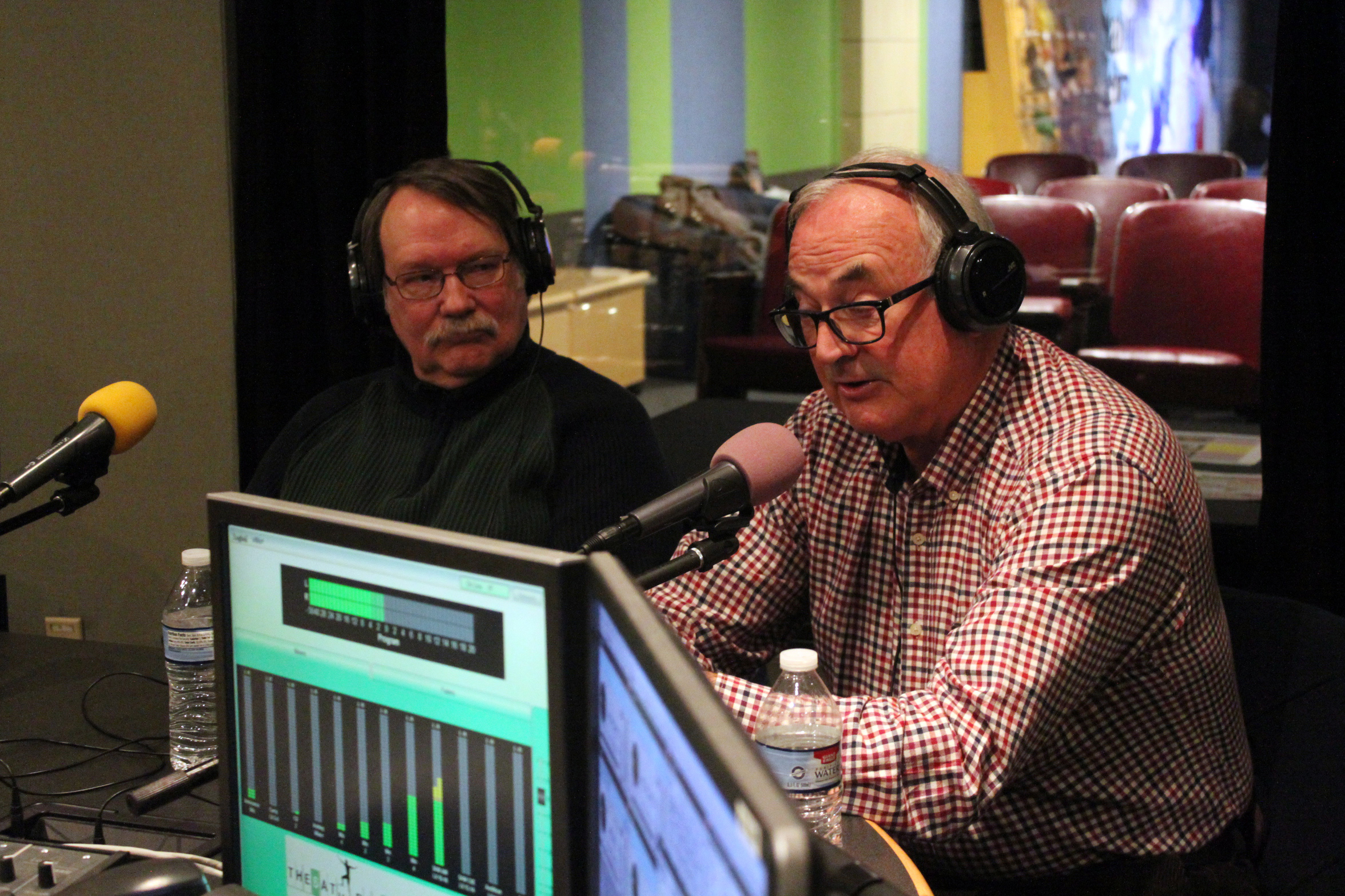 Two adults wearing headphones speaking into microphones in a radio studio.