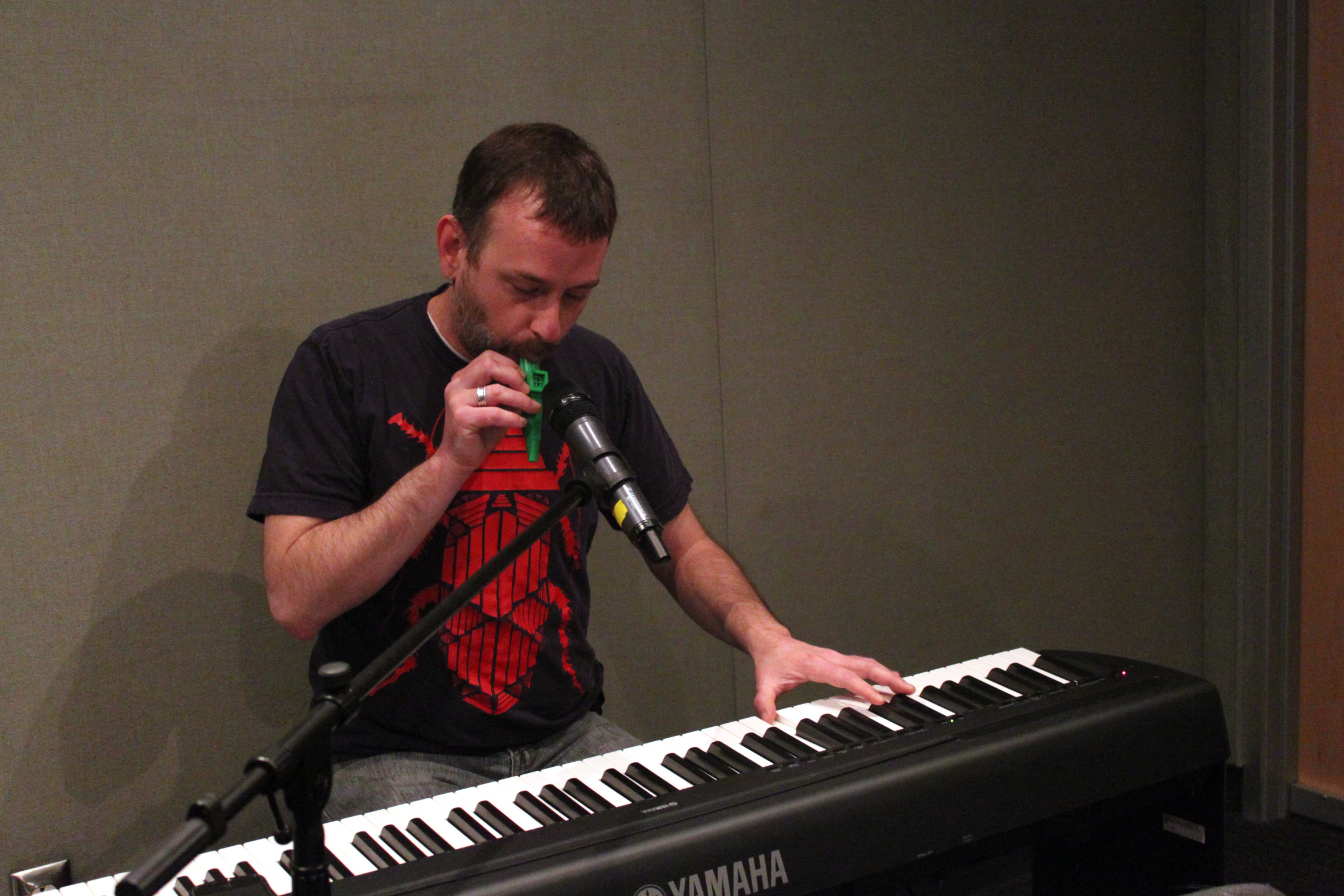 An adult playing a kazoo into a microphone while playing a piano keyboard in a radio studio