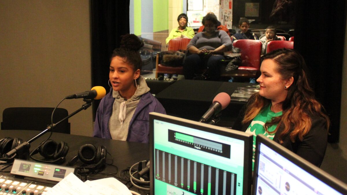 Two teens speaking into headphones in a radio studio