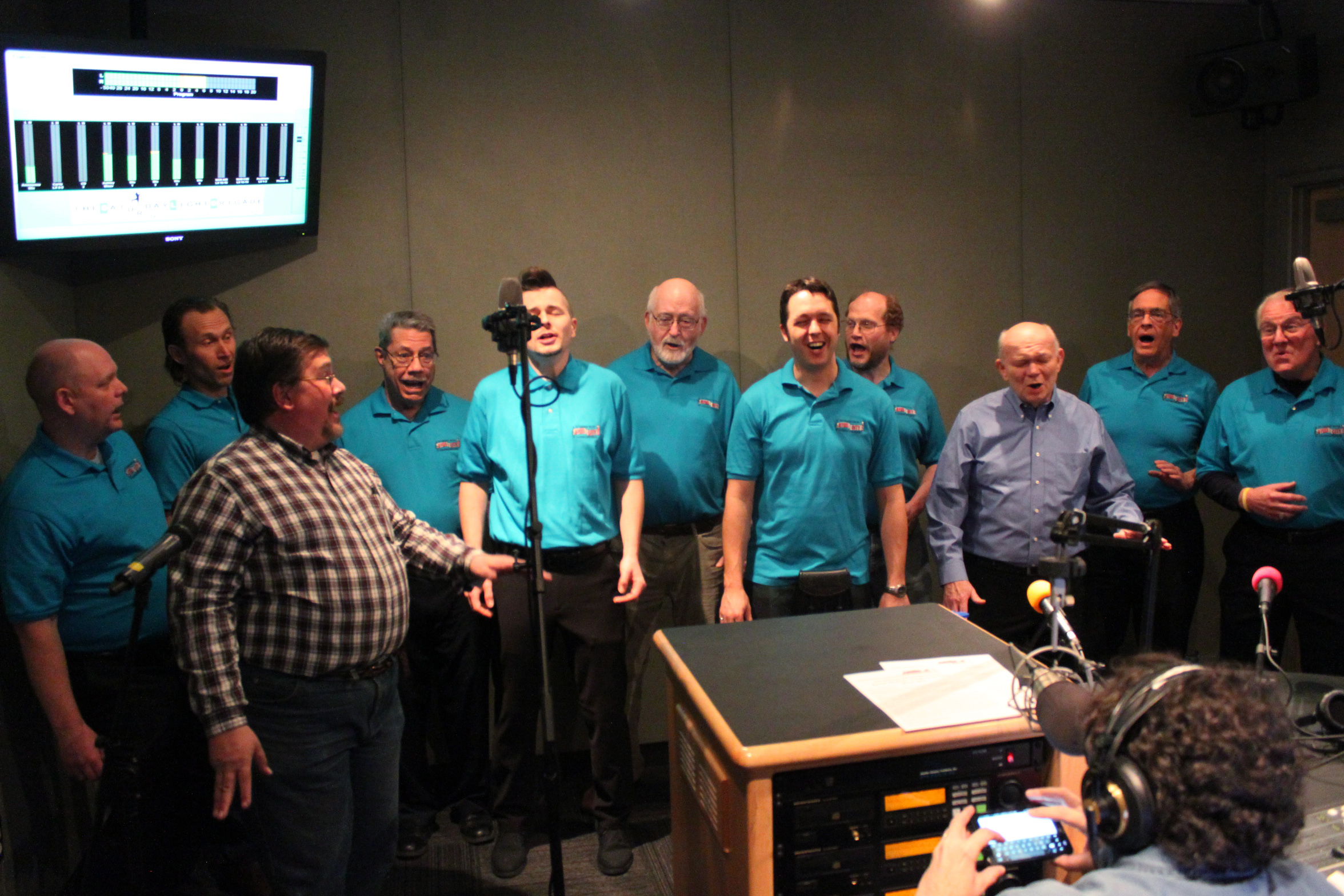 Eleven adults singing as a choir into microphones as another adult wearing headphones listens in a radio studio