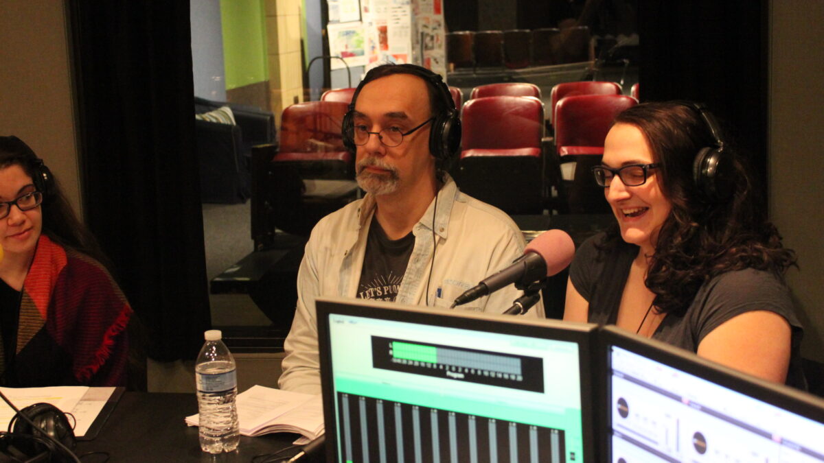 Three adults wearing headphones speaking into microphones in a radio studio