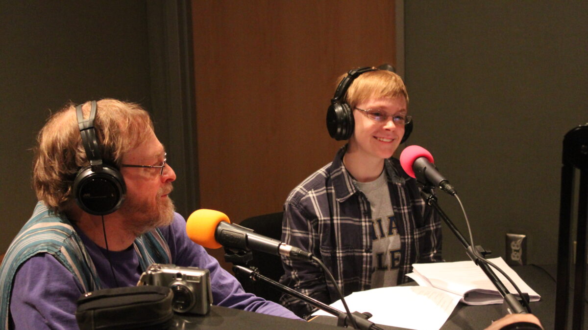 Two adults wearing headphones speaking into microphones in a radio studio