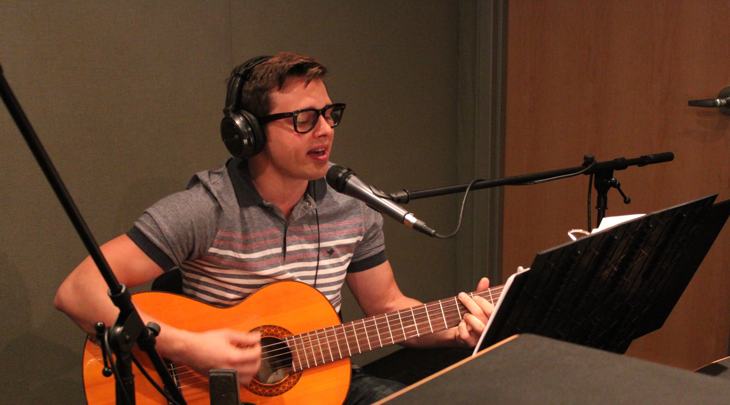 An adult wearing headphones singing into a microphone and playing acoustic guitar in a radio studio