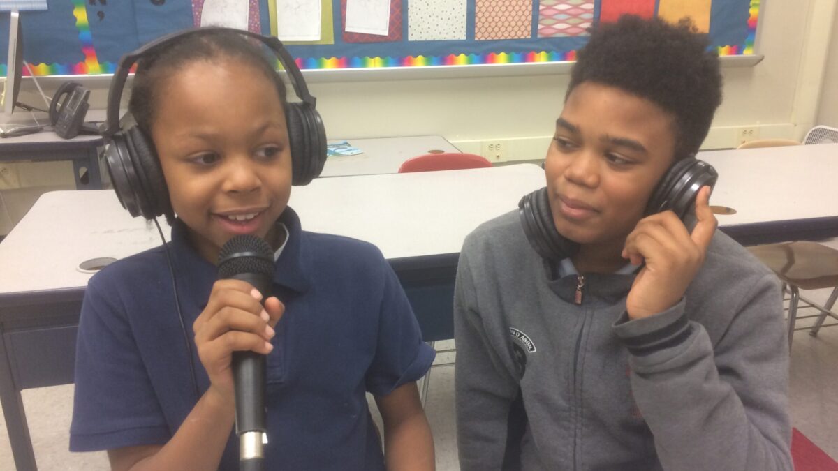 Two youth wearing headphones in a classroom, one holding and speaking into a microphone