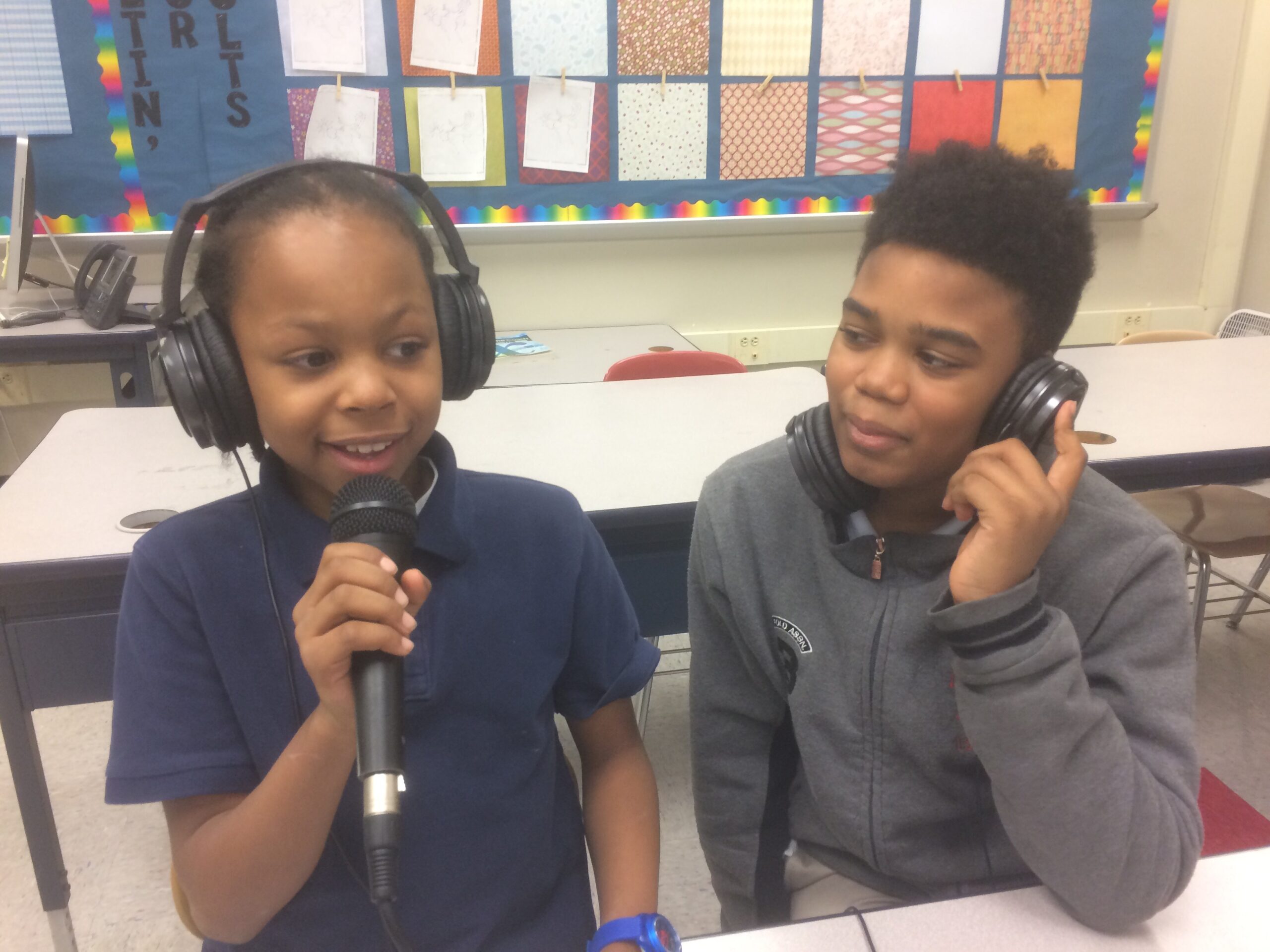 Two youth wearing headphones in a classroom, one holding and speaking into a microphone