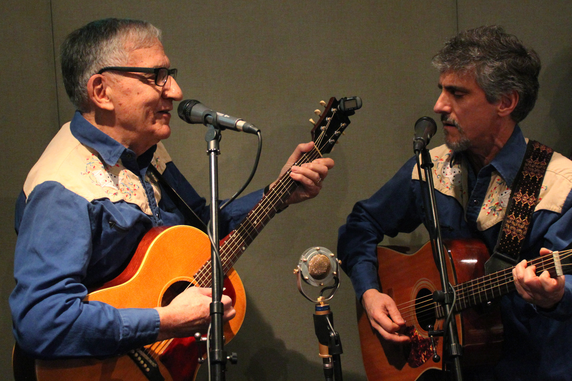 Two adults playing acoustic guitars and singing into microphones in a radio studio