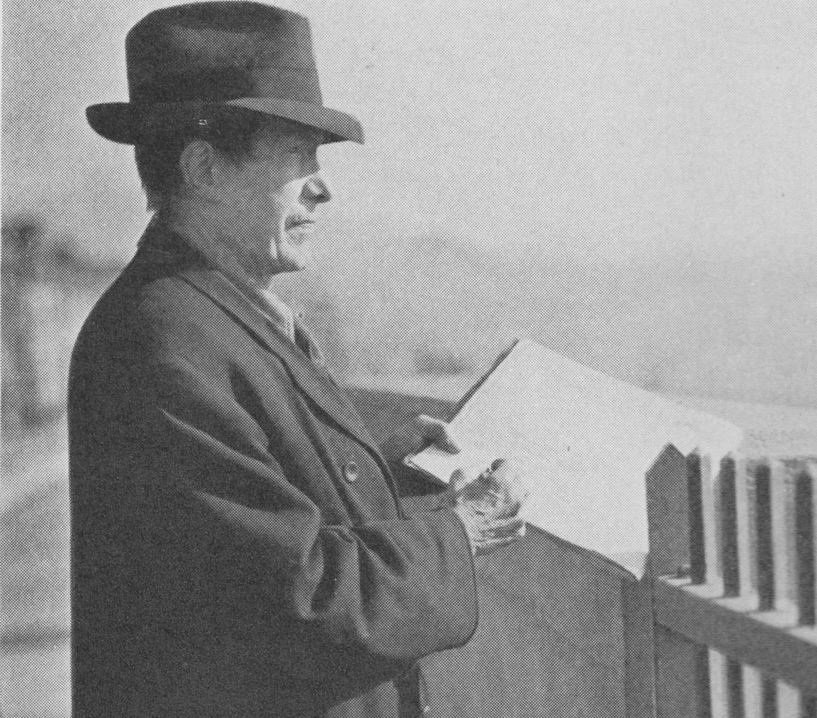 A black and white photo of an individual sketching on a piece of paper while looking over a fence