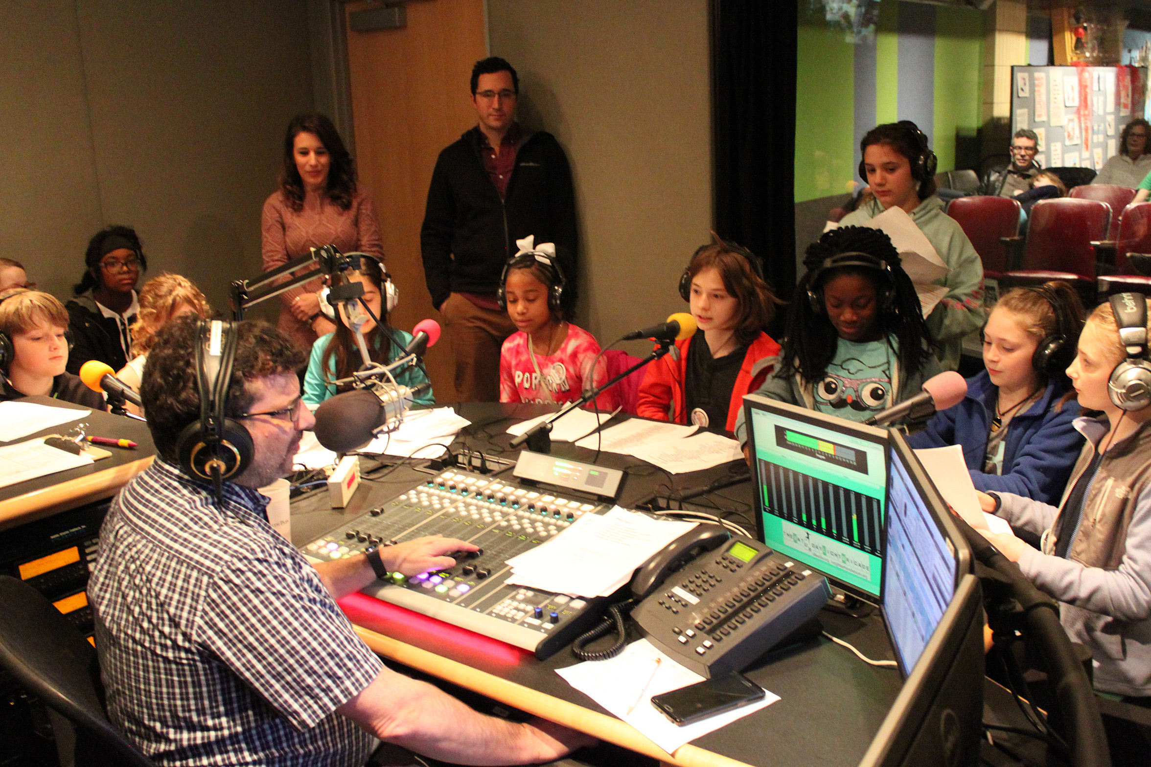 Nine youth wearing headphones sitting behind microphones being interviewed by an adult wearing headphones sitting behind a microphone and a mixing board in a radio studio as four adults listen in the background