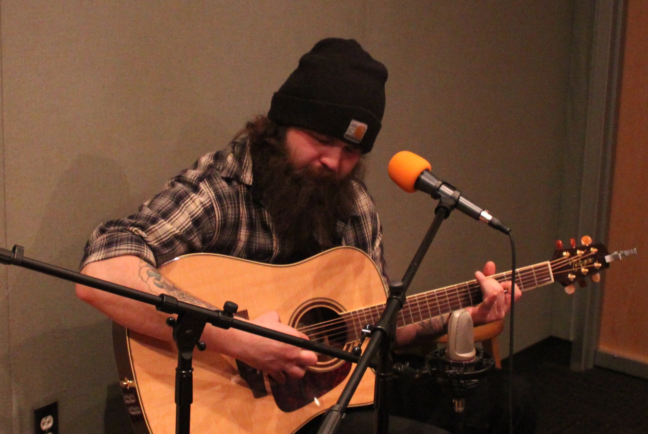 An adult playing acoustic guitar sitting behind a microphone in a radio studio