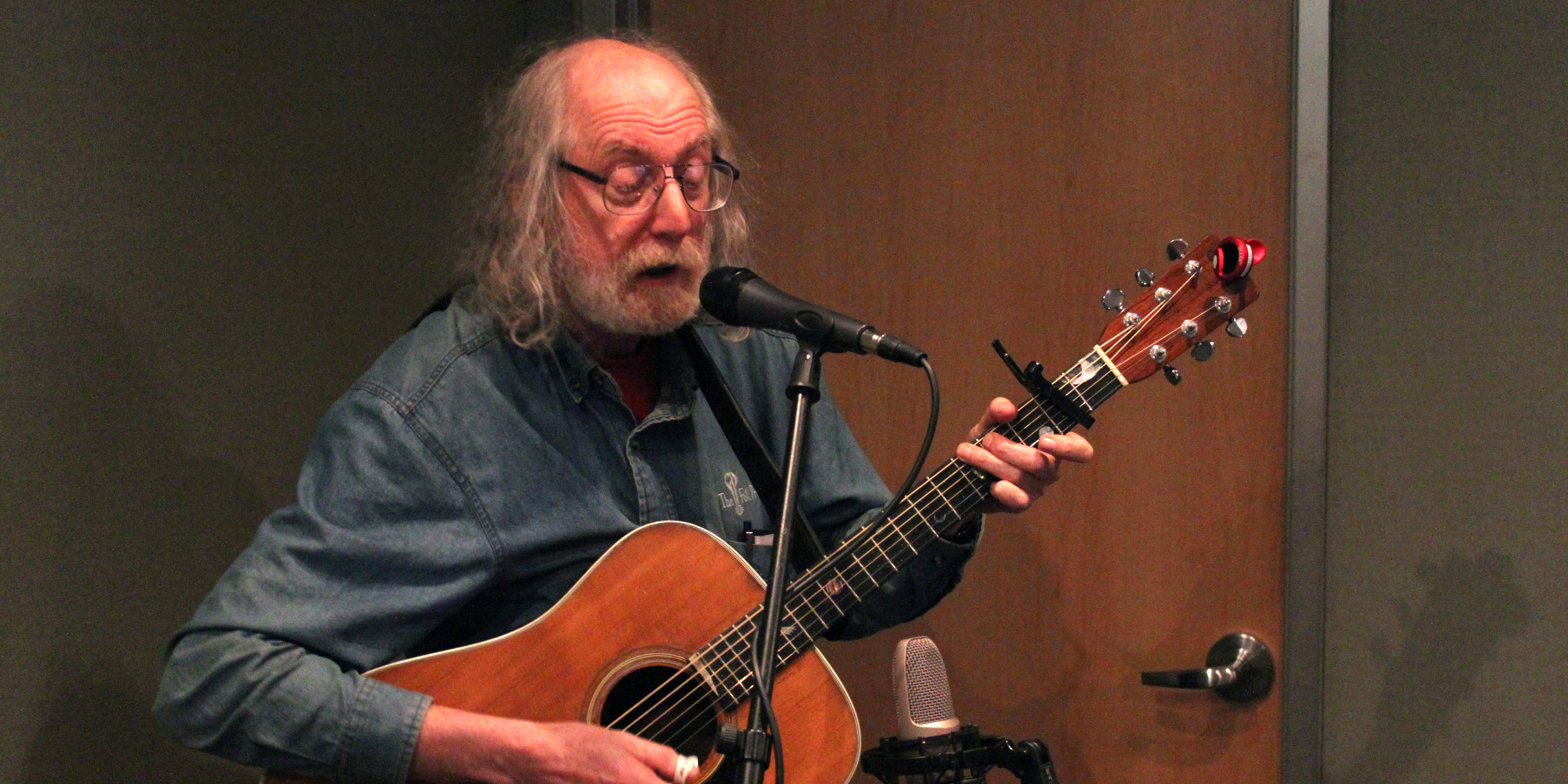 An adult playing acoustic guitar singing into a microphone in a radio studio