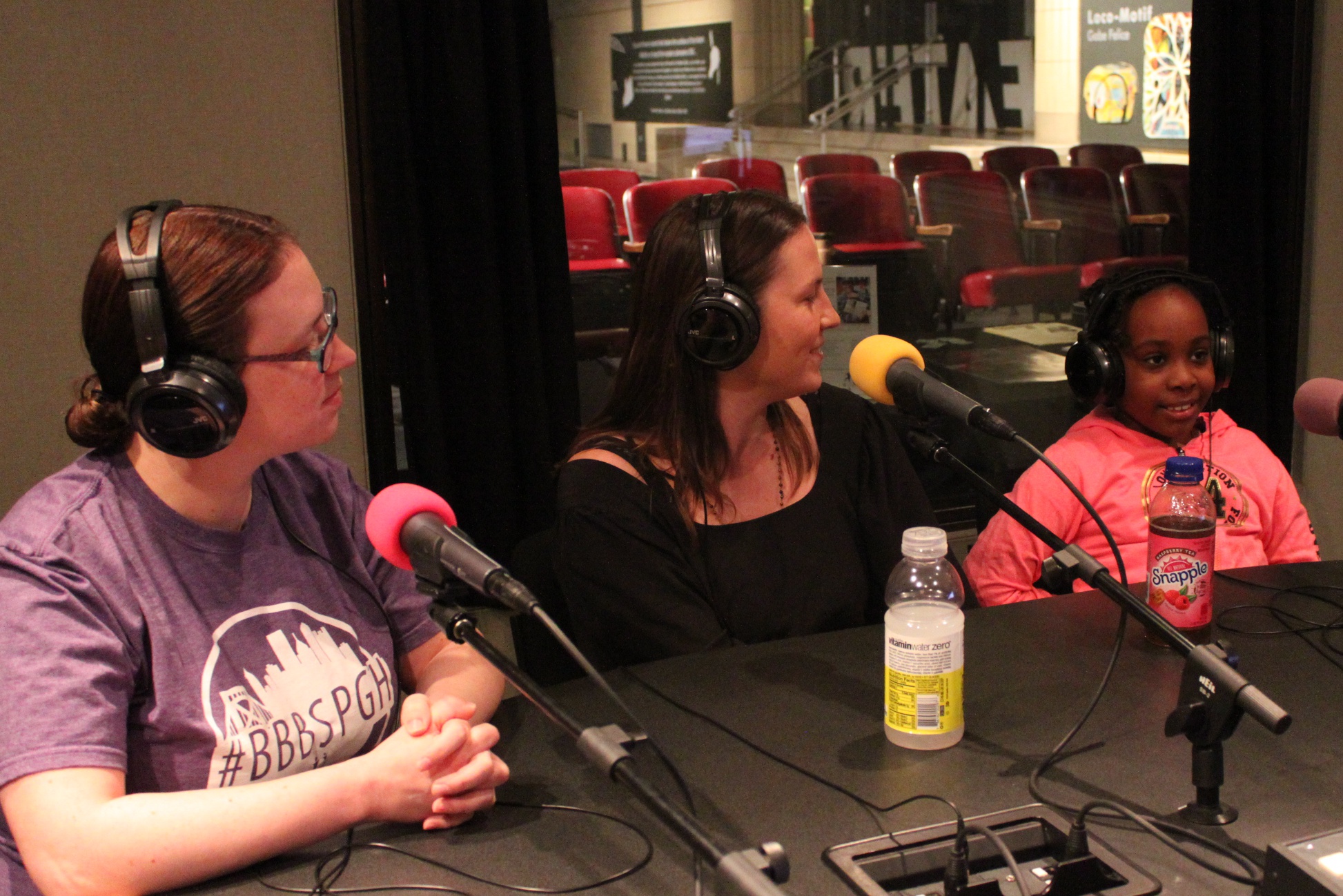 Two adults and a youth wearing headphones speaking into microphones in a radio studio