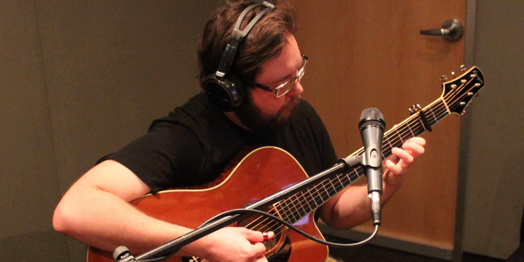 An adult wearing headphones playing acoustic guitar and singing into a microphone in a radio studio