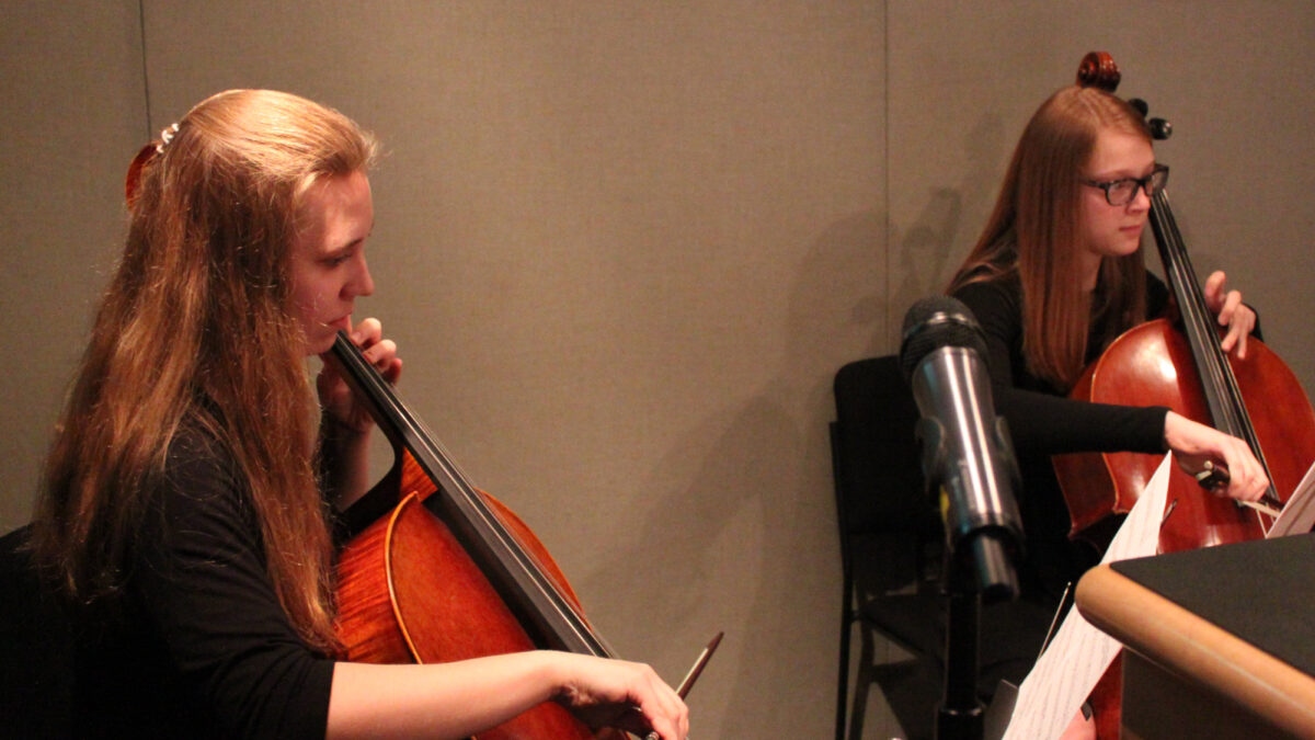 Two teens playing cello looking at sheet music on music stands in a radio studio