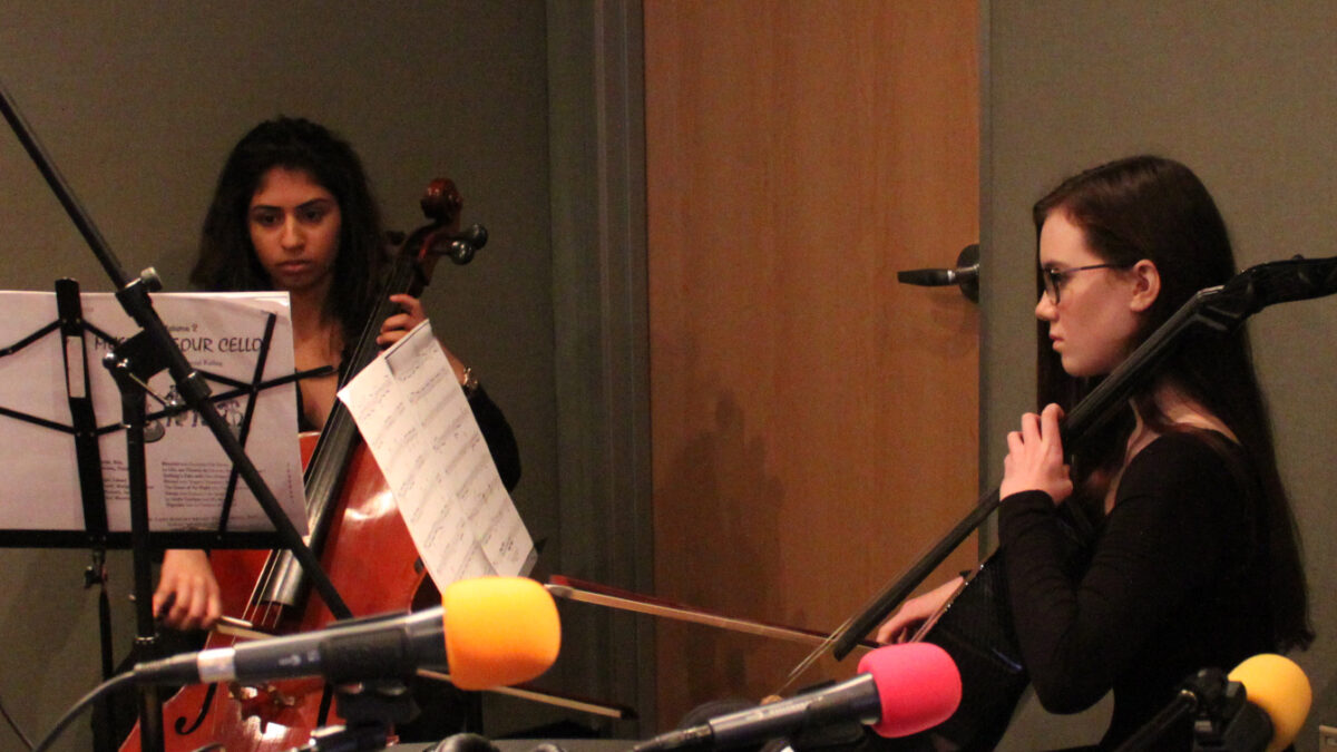 Two teens playing cello looking at sheet music on music stands in a radio studio