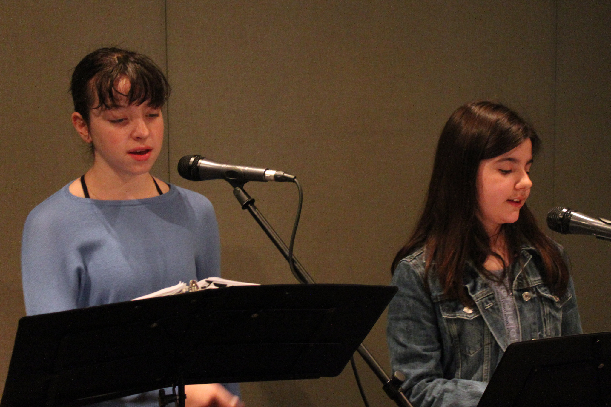 Two youth singing into microphones looking at sheet music on music stands in a radio studio