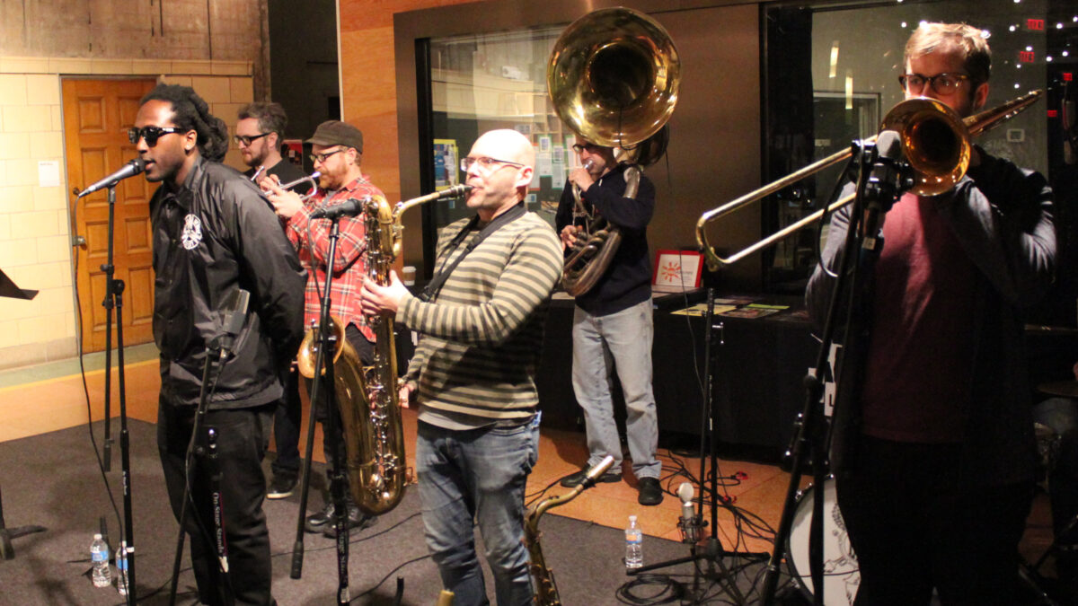 Two adults playing trumpet, an adult singing into a microphone, an adult playing baritone saxophone, an adult playing sousaphone, and an adult playing trombone