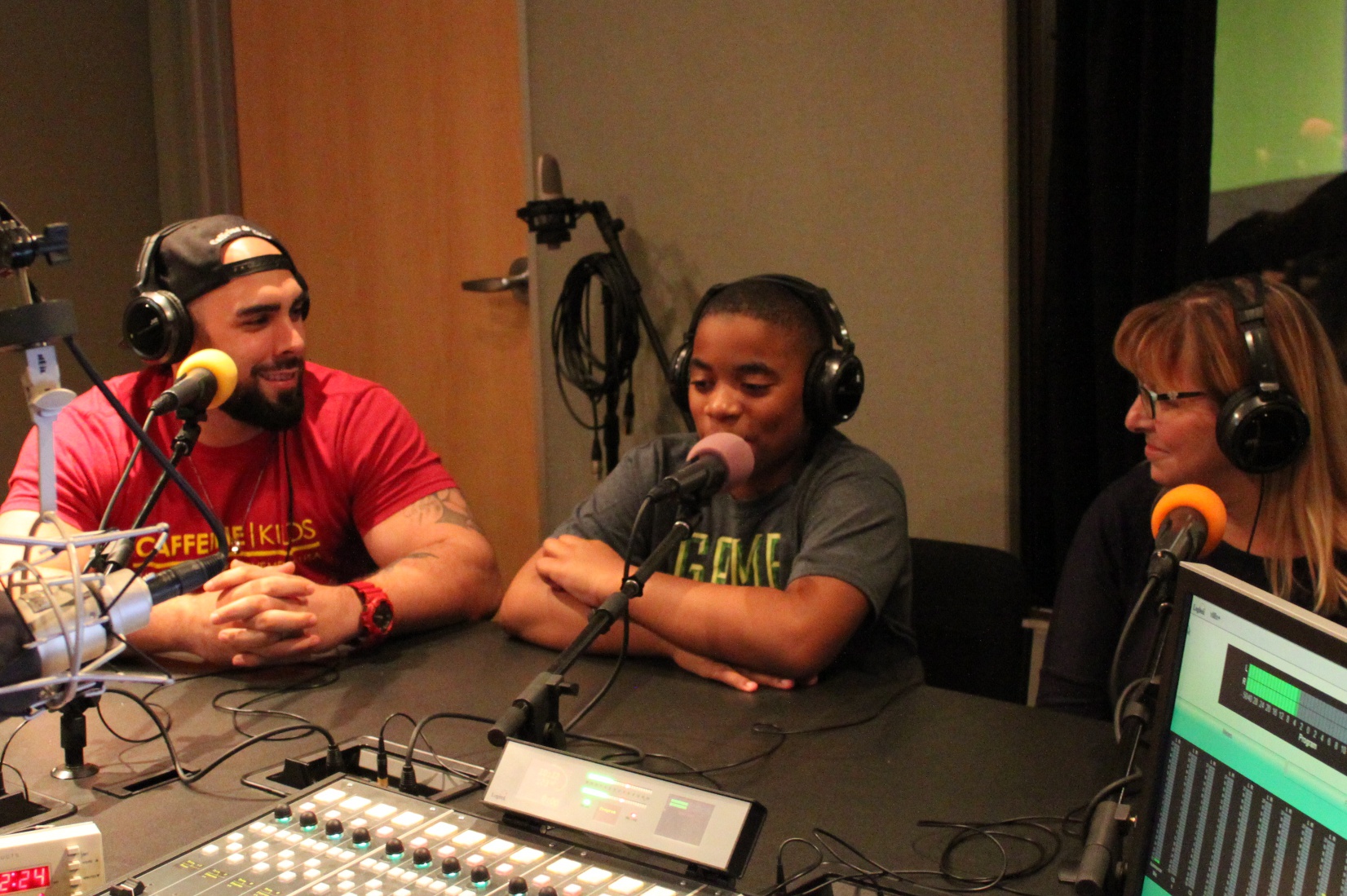 Two adults and a youth wearing headphones speaking into microphones in a radio studio