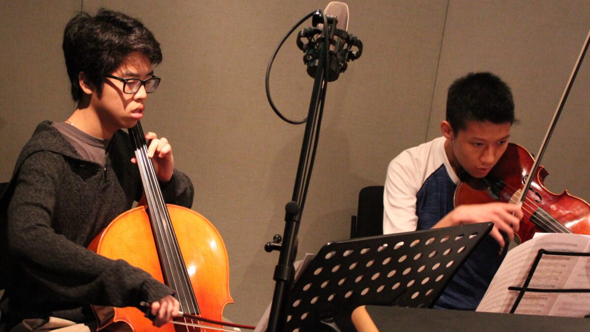 A teen playing cello and a teen playing viola looking at sheet music on music stands in a radio studio