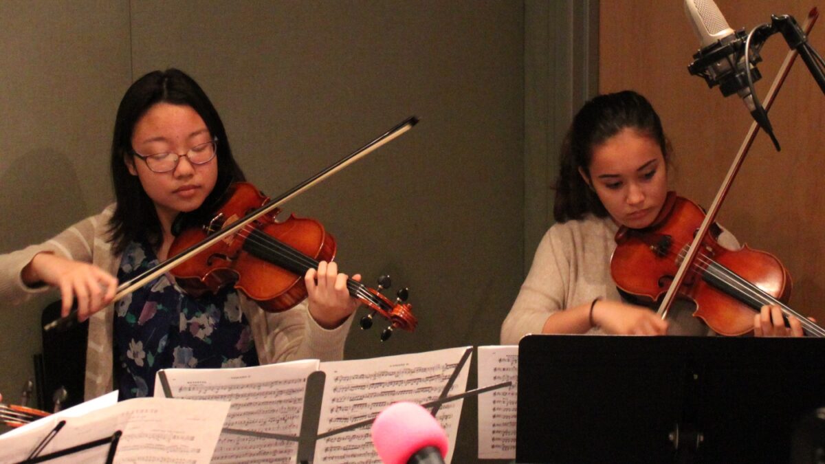 Two teens playing violins looking at sheet music on music stands in a radio studio