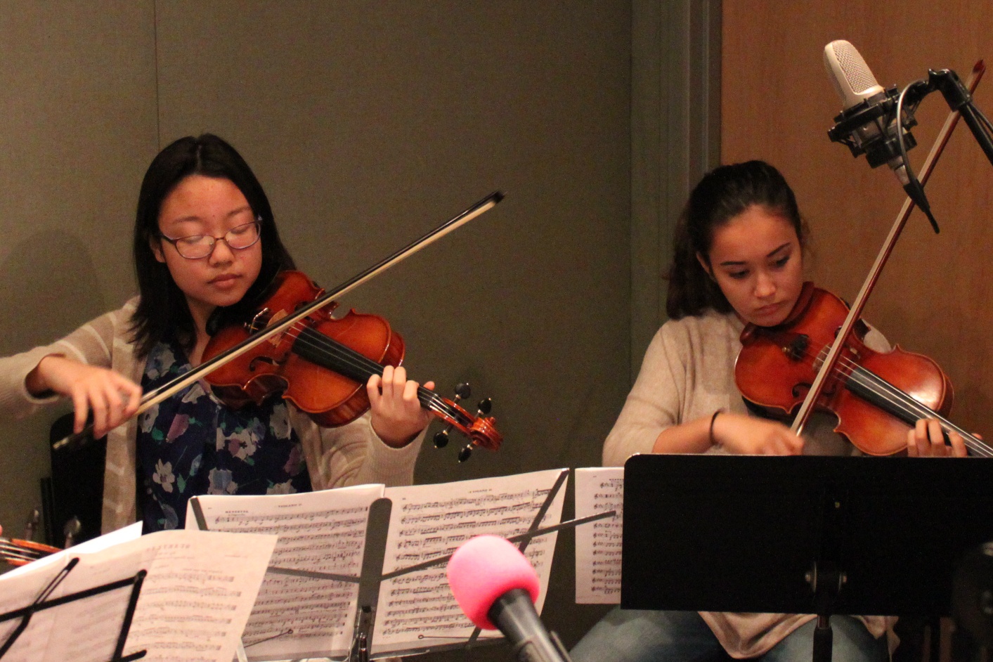 Two teens playing violins looking at sheet music on music stands in a radio studio