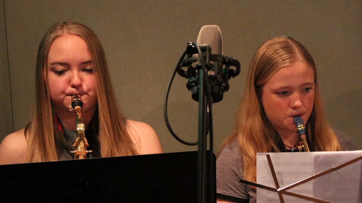 Two teens playing alto saxophones looking at sheet music on music stands in a radio studio