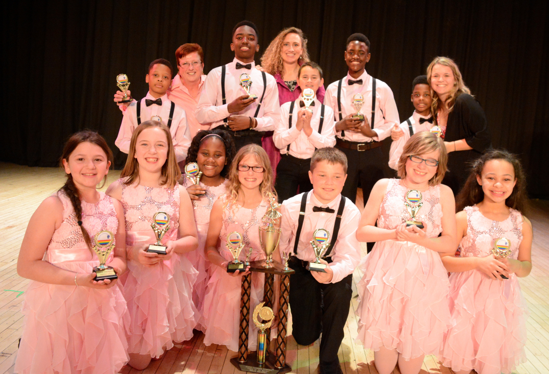 Twelve youth and three adults smiling and posing behind a gold trophy