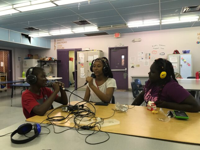Three youth wearing headphones speaking into microphones connected to a portable recorder in a classroom