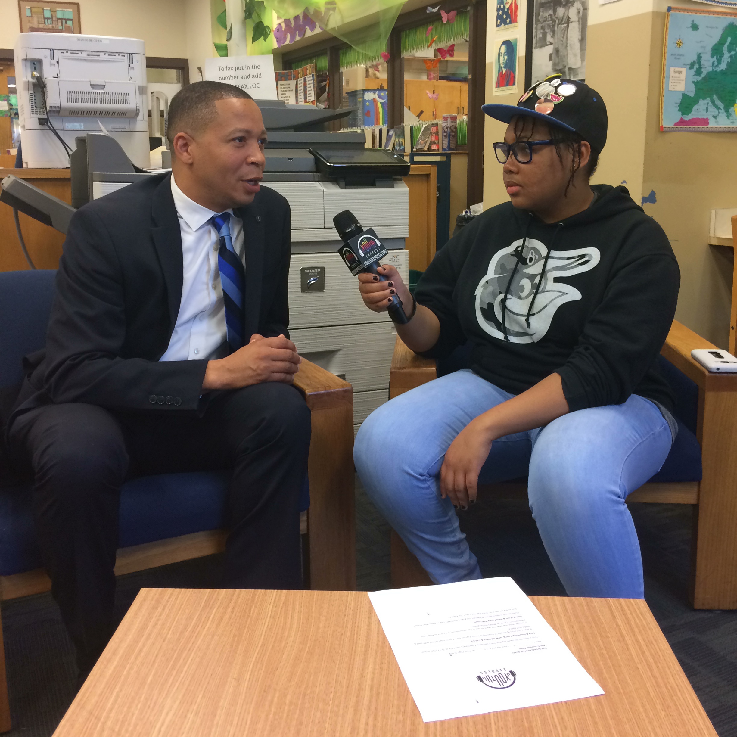 A teen holding a microphone interviewing an adult wearing a suit in an office