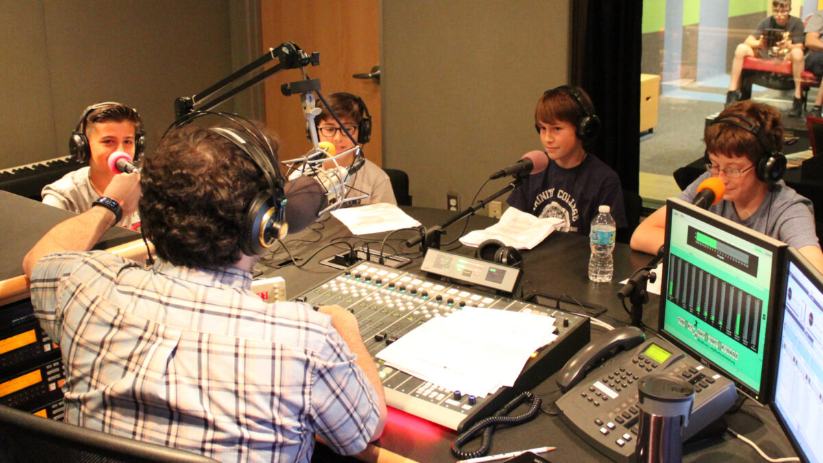 Four youth wearing headphones sitting behind microphones being interviewed by an adult wearing headphones sitting behind a microphone and a mixing board in a radio studio