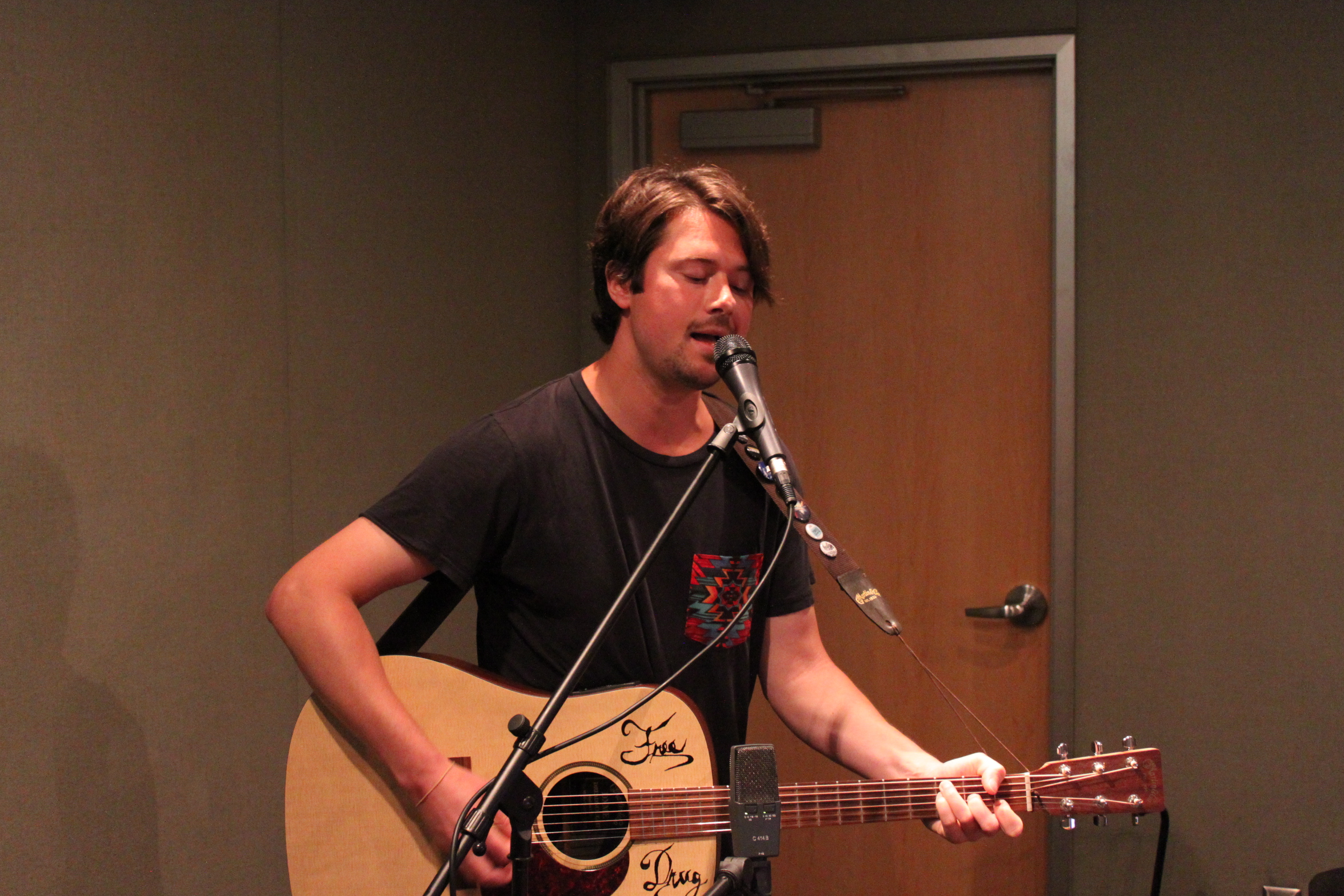 An adult playing acoustic guitar and singing into a microphone in a radio studio