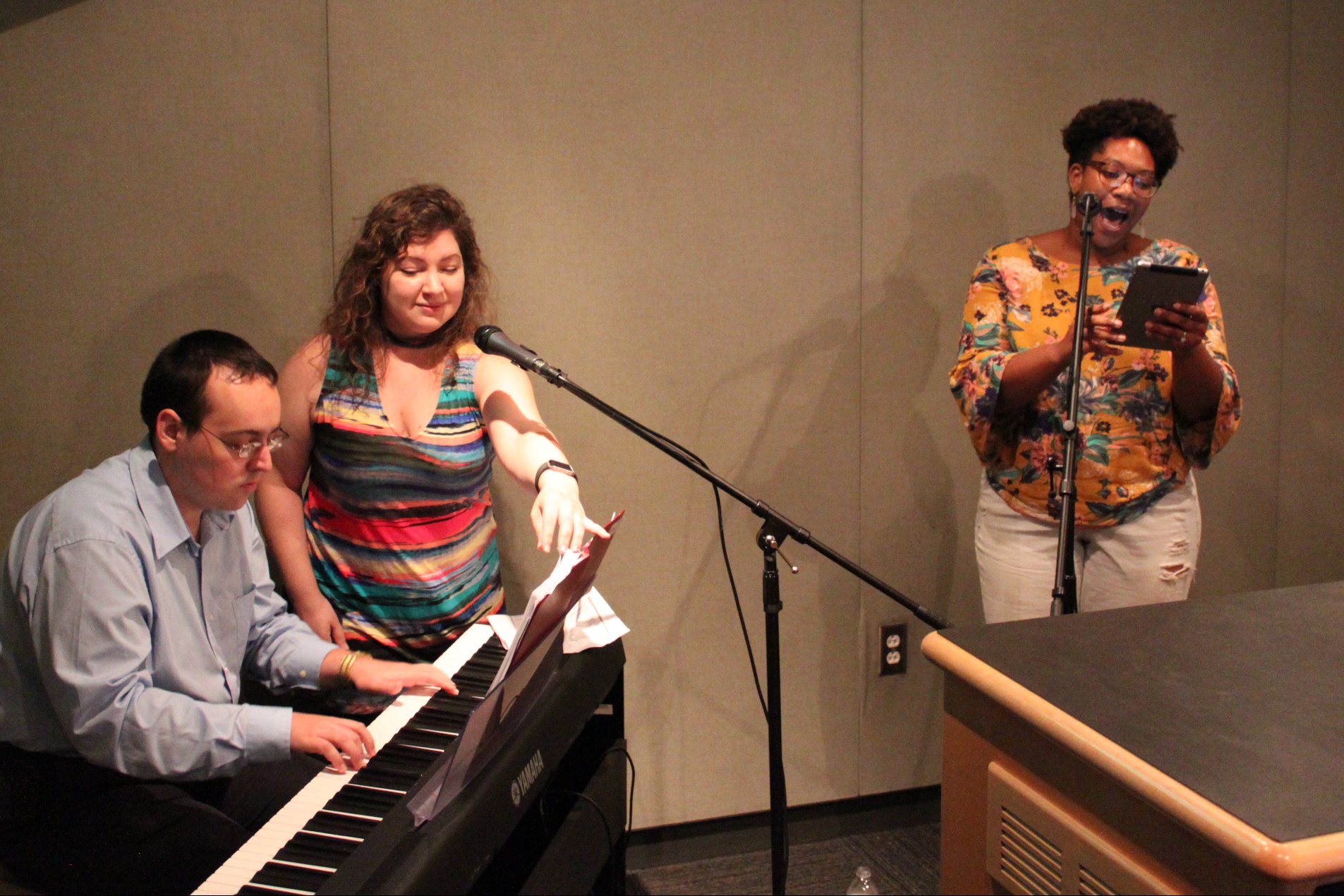 An adult playing a piano keyboard and two adults singing into microphones in a radio studio
