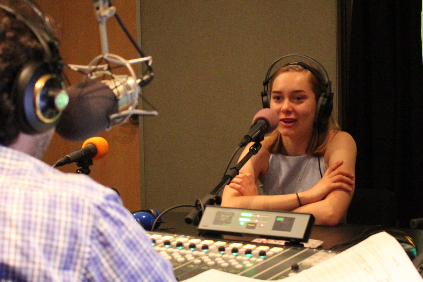 A teen wearing headphones speaking into a microphone being interviewed by an individual sitting behind a microphone and a mixing board in a radio studio