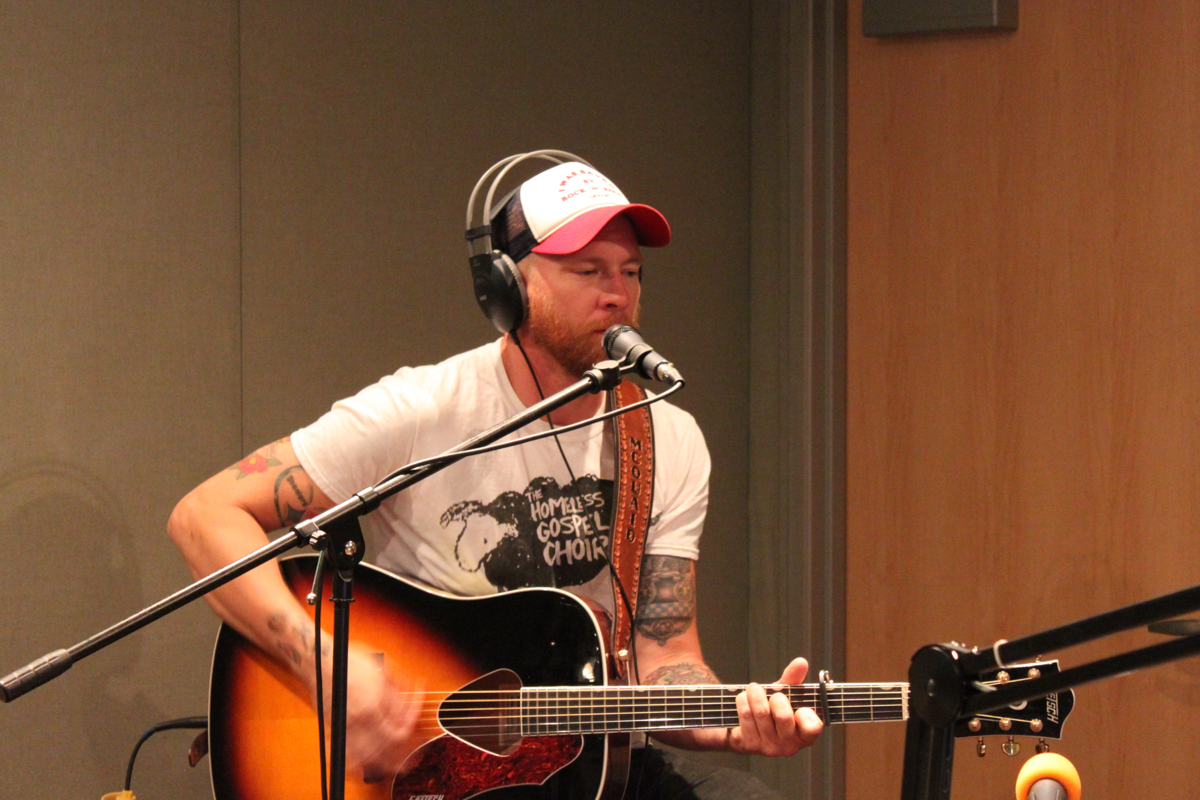 An adult wearing headphones playing acoustic guitar and singing into a microphone in a radio studio