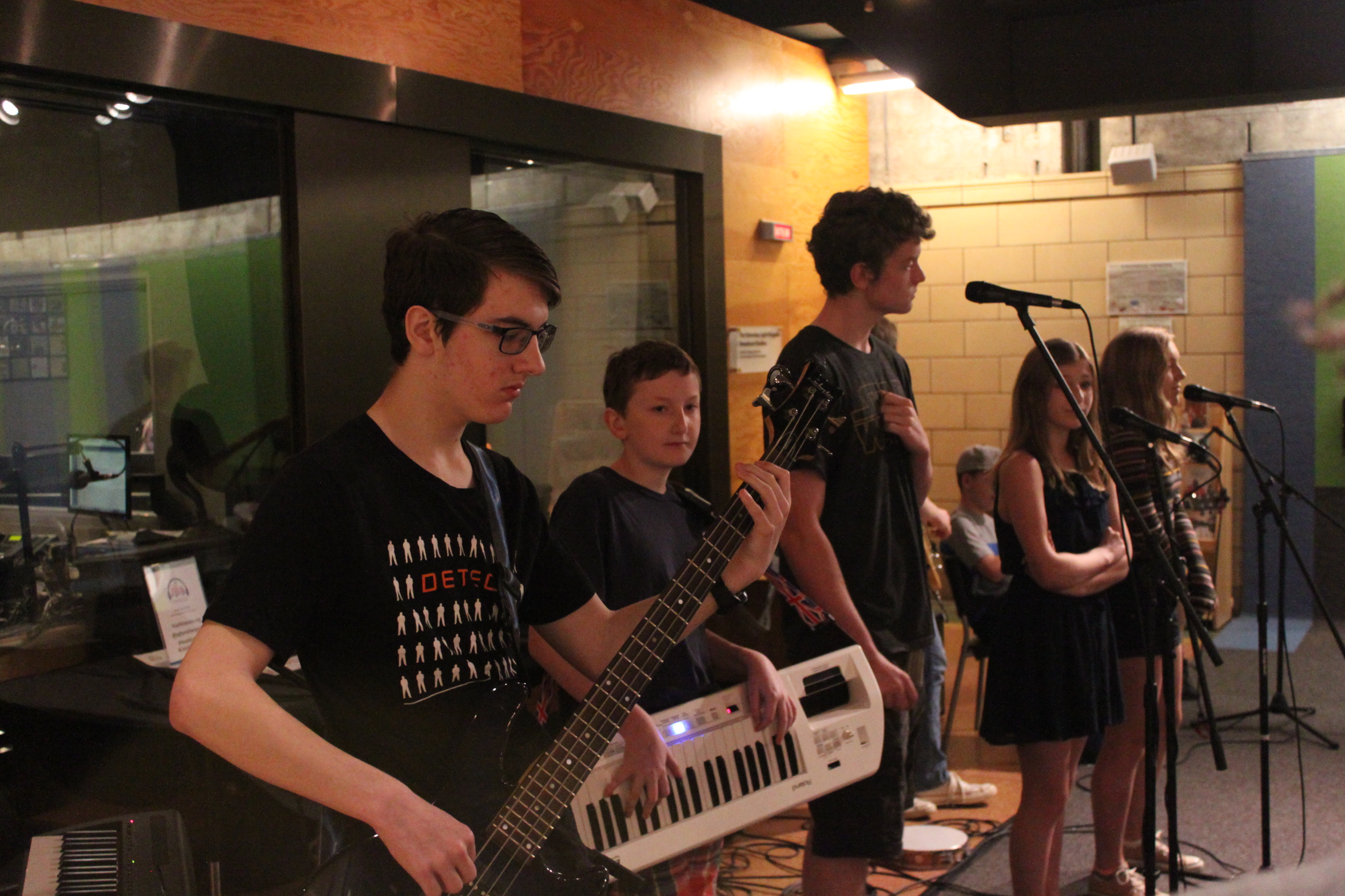 A teen playing electric bass, a youth playing keytar, and three youth singing into microphones