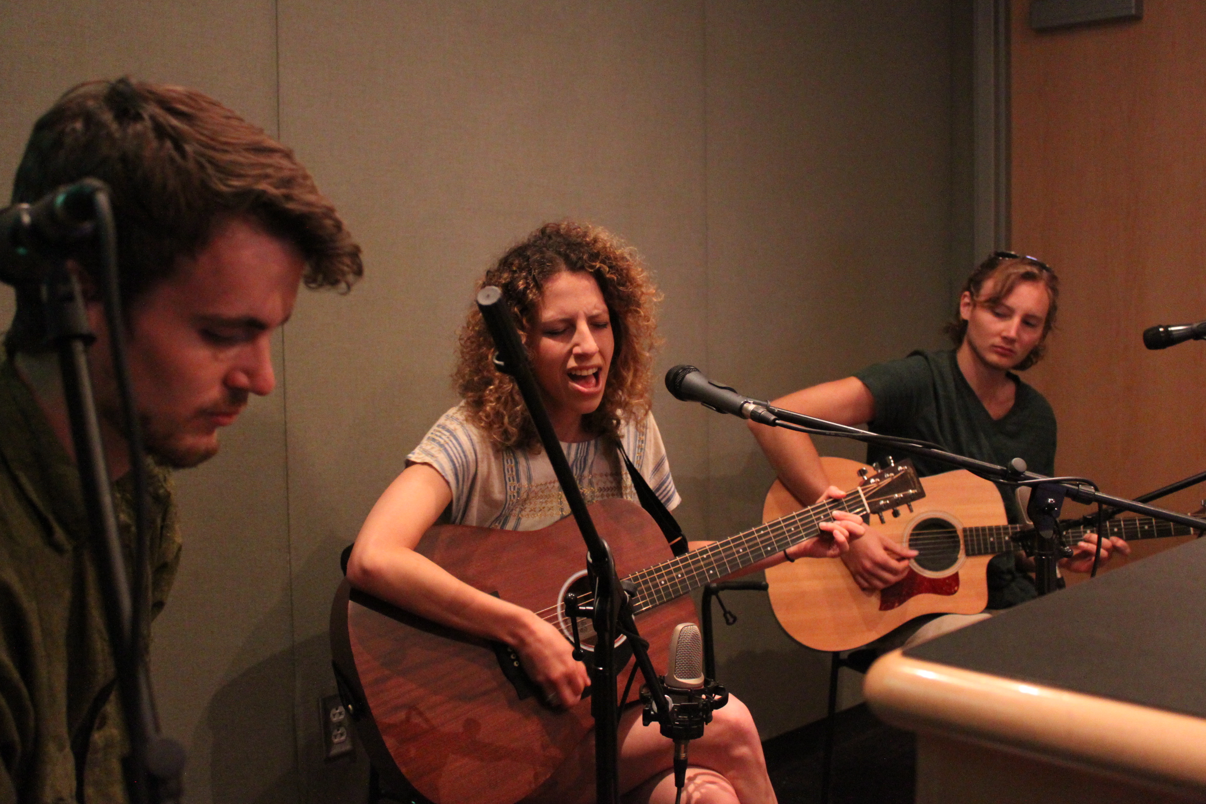 Two adults playing acoustic guitar and singing into microphones, and an adult sitting behind a microphone in a radio studio