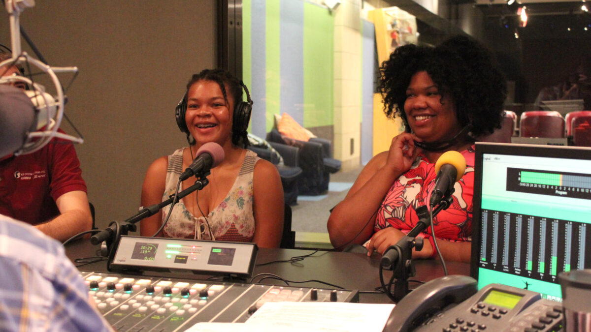 Two teens wearing headphones laughing into microphones in a radio studio