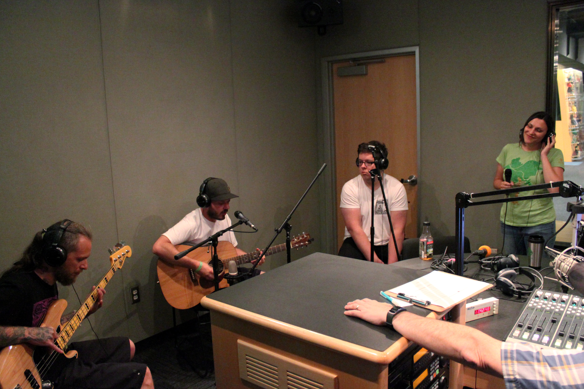 An adult playing electric bass, an adult playing acoustic guitar, and two adults singing into microphones, all wearing headphones in a radio studio