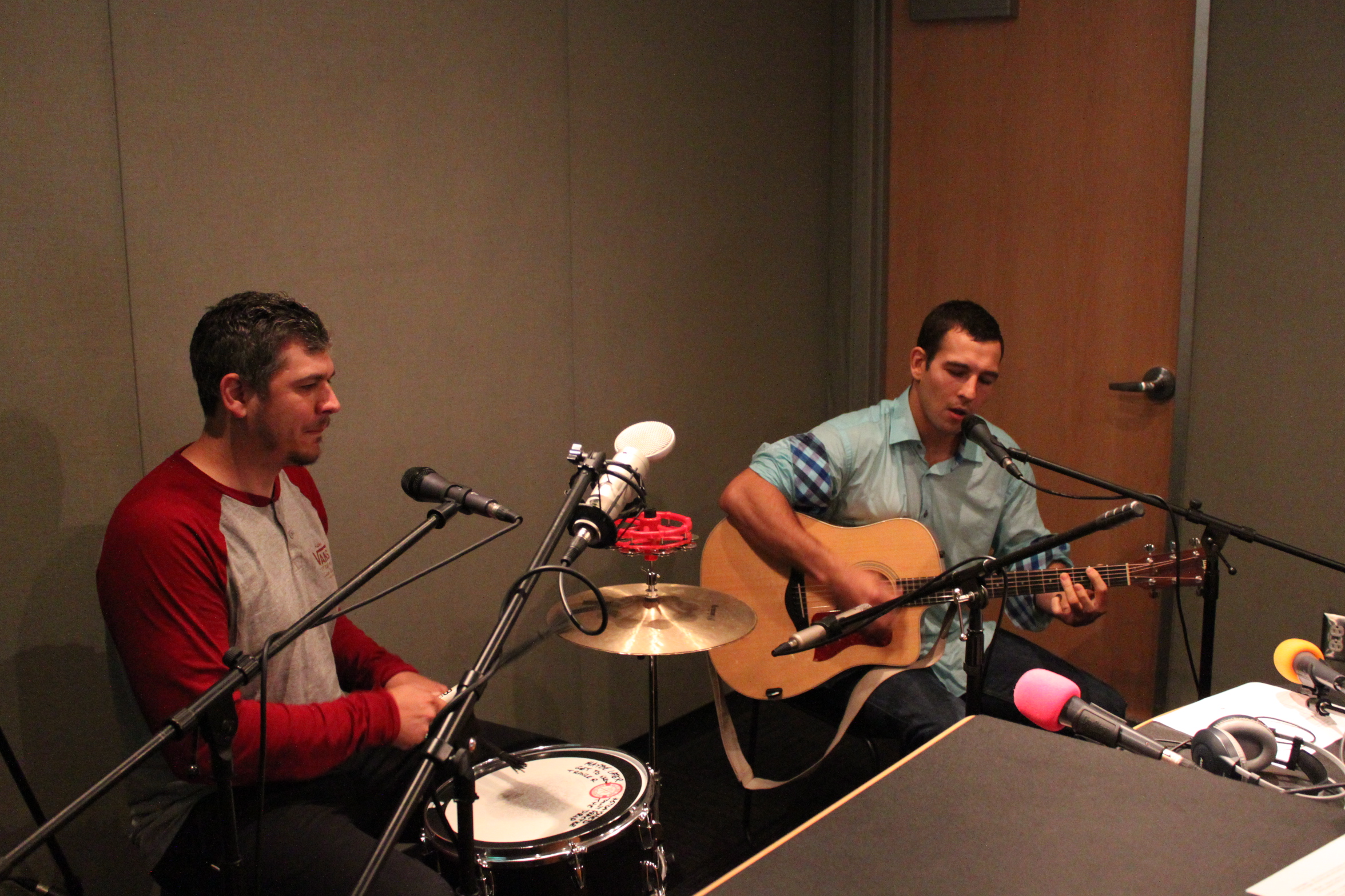 An adult playing a small drum set and an adult playing acoustic guitar and singing into a microphone in a radio studio