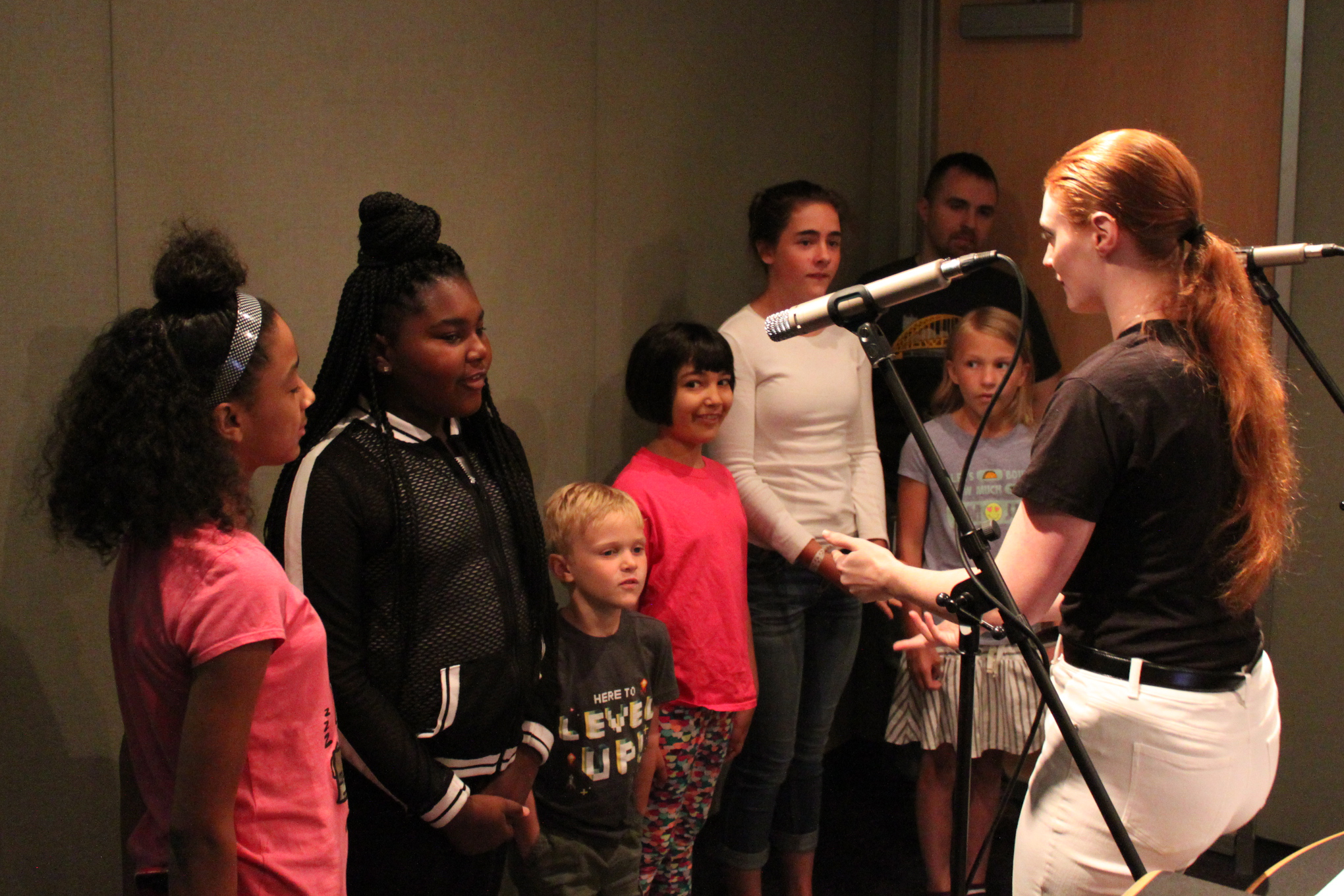 Six youth singing in a choir being led by an adult singing into a microphone in a radio studio