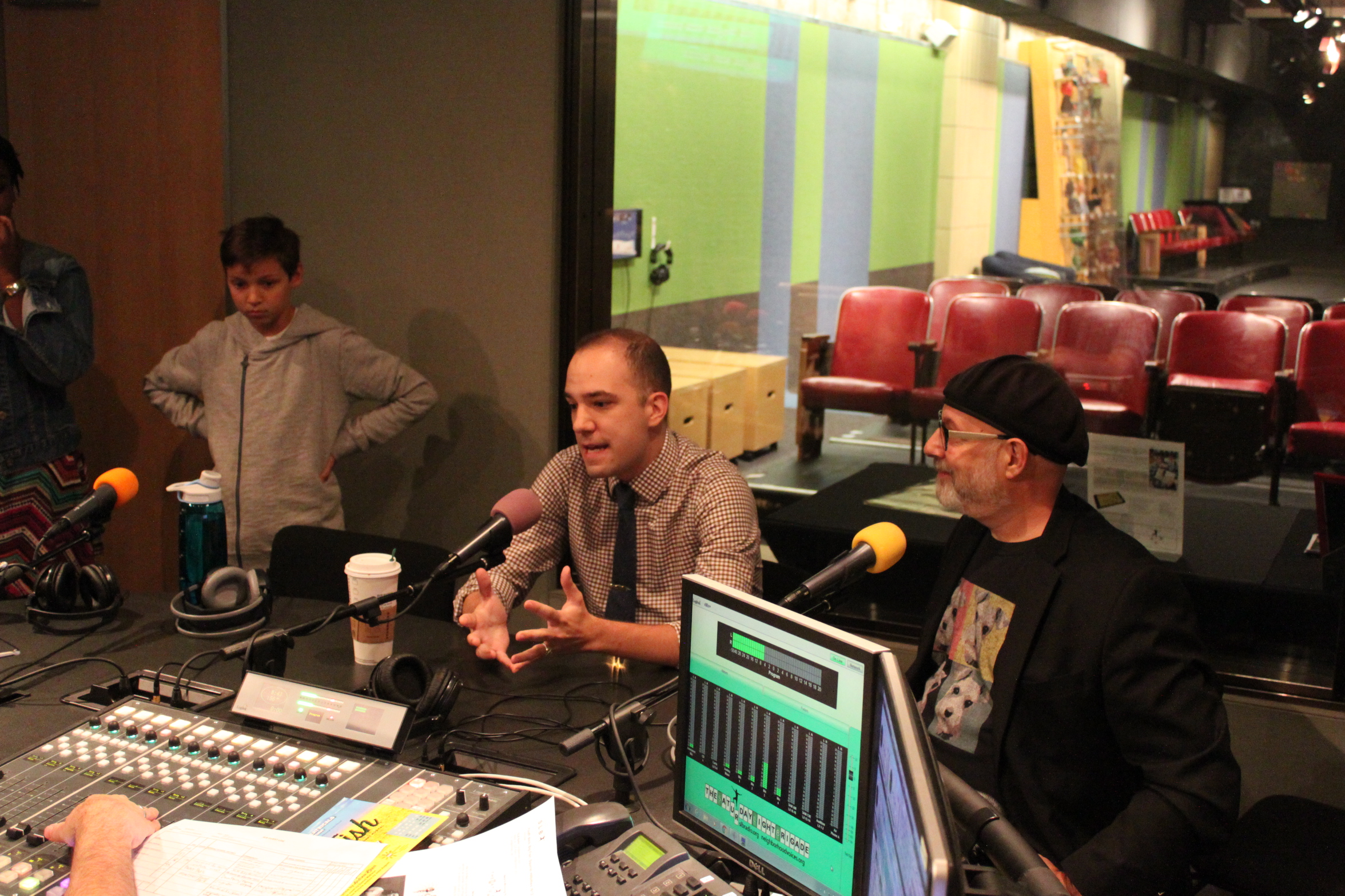 Two adults sitting and speaking into microphones and one child standing nearby in a radio studio