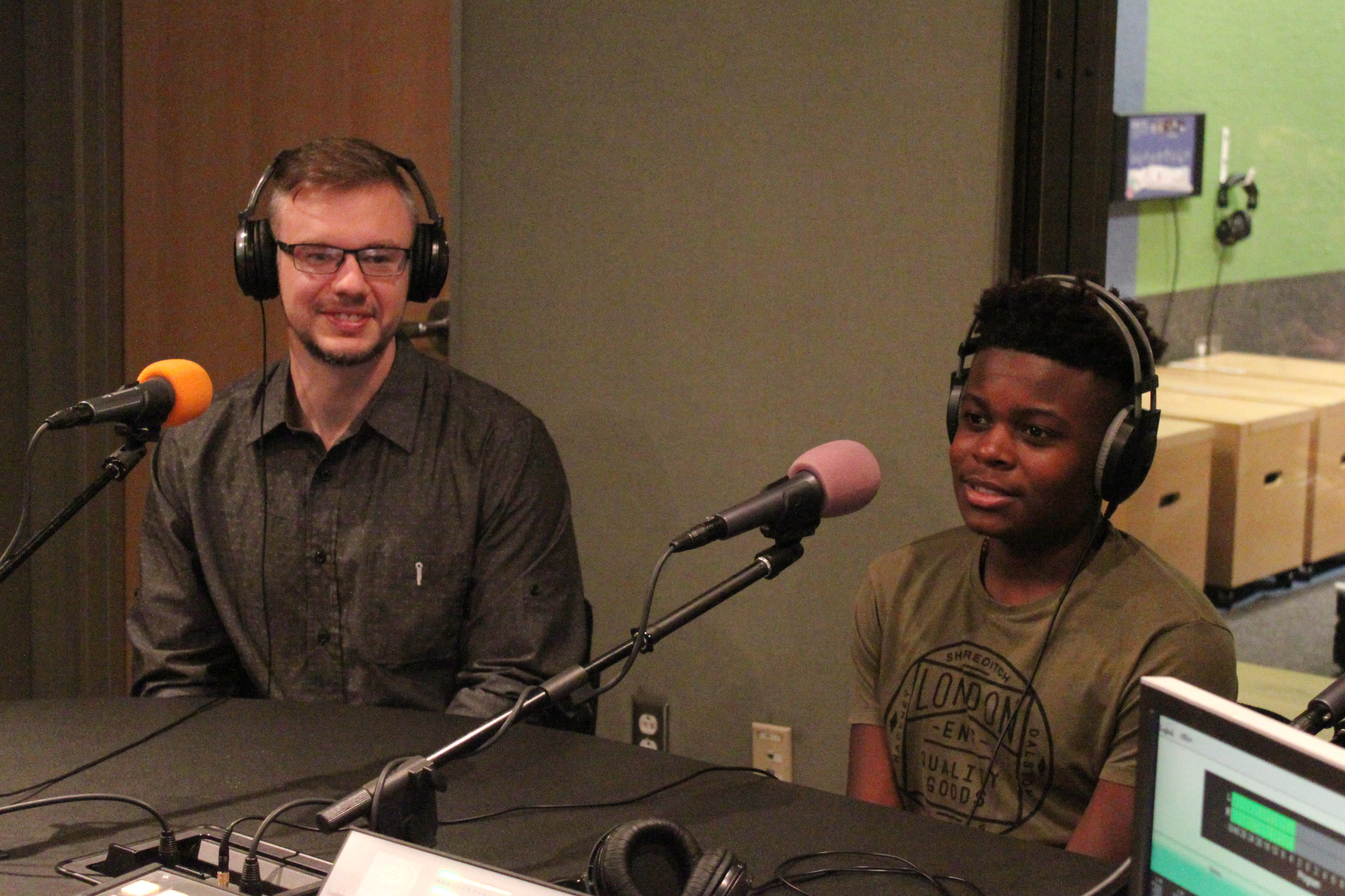 An adult and a teen wearing headphones sitting behind microphones in a radio studio