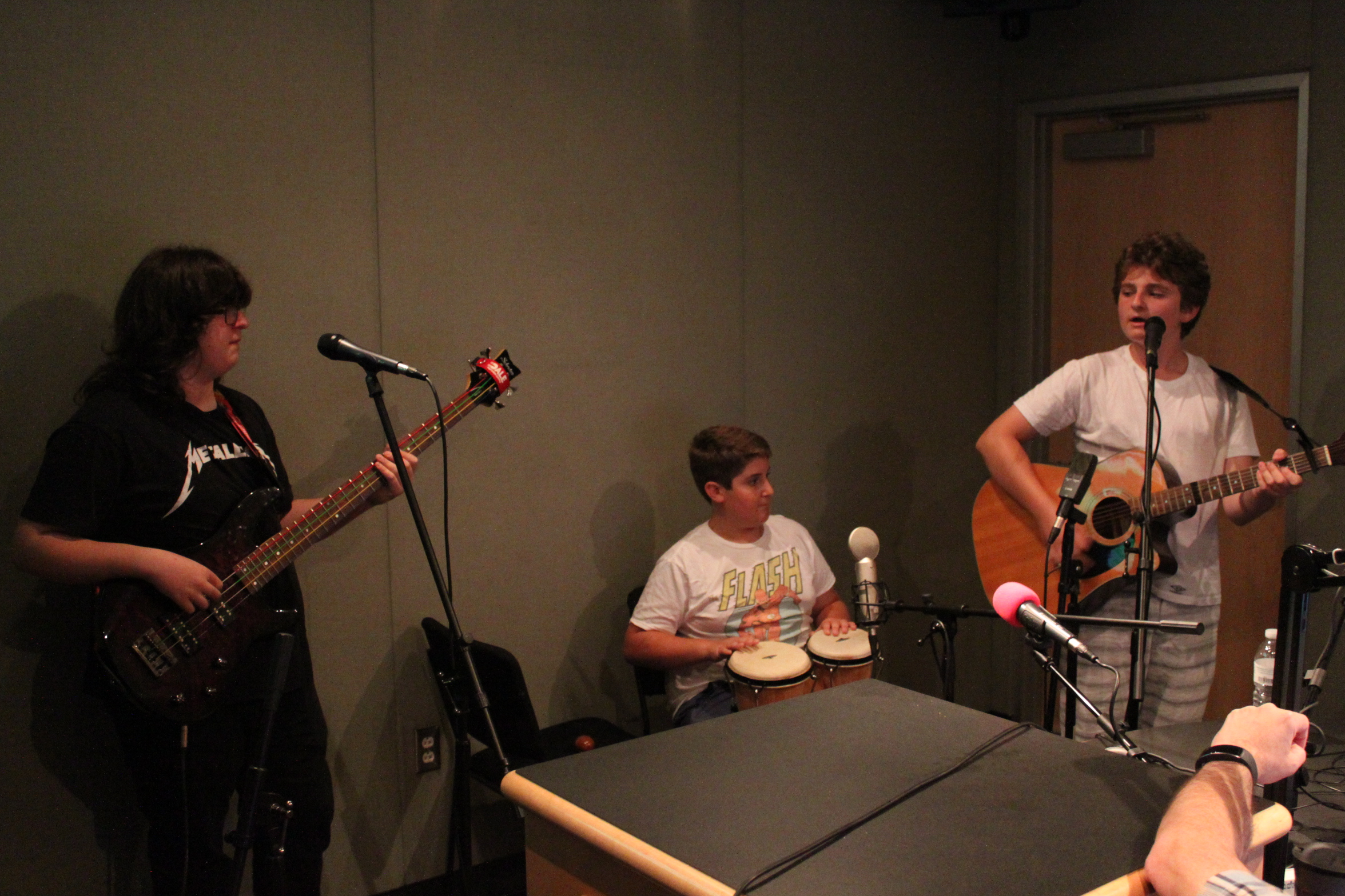 A teen playing electric bass, a teen playing congas, and a teen playing acoustic guitar in a radio studio